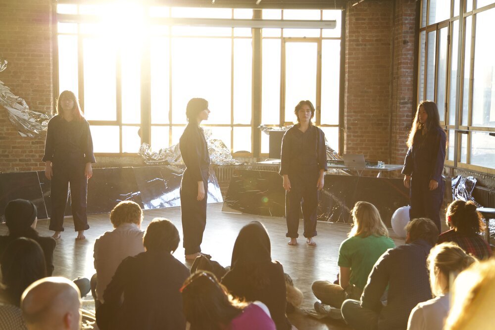 four actors in workmen jumpers soaked in light from large windows behind them. they are in front of an audience