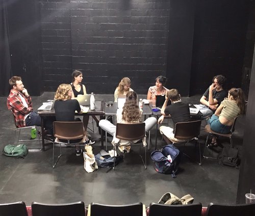 nine people sitting around a table in the middle of a stage, doing a tableread