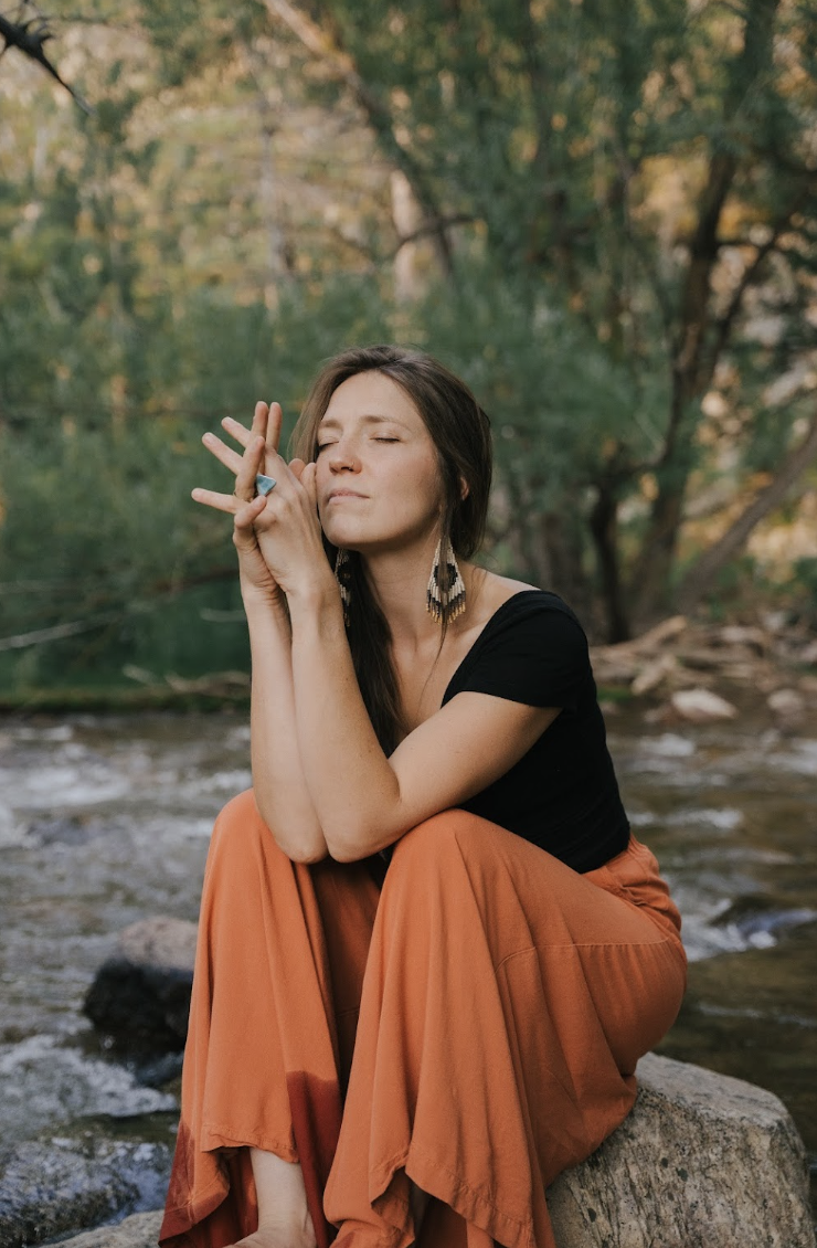 A woman with long brown hair, wearing a black top, orange pants, and feather earrings, sitting on a rock by a creek with trees in the background, eyes closed and hands clasped near her face.