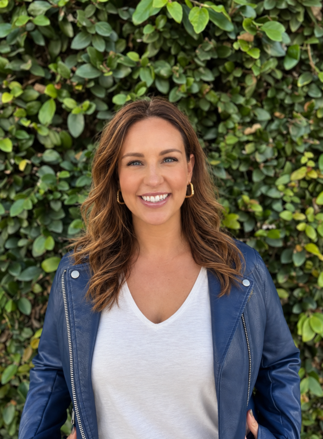 A smiling woman in a white shirt and blue jacket standing in front of a green leafy hedge.