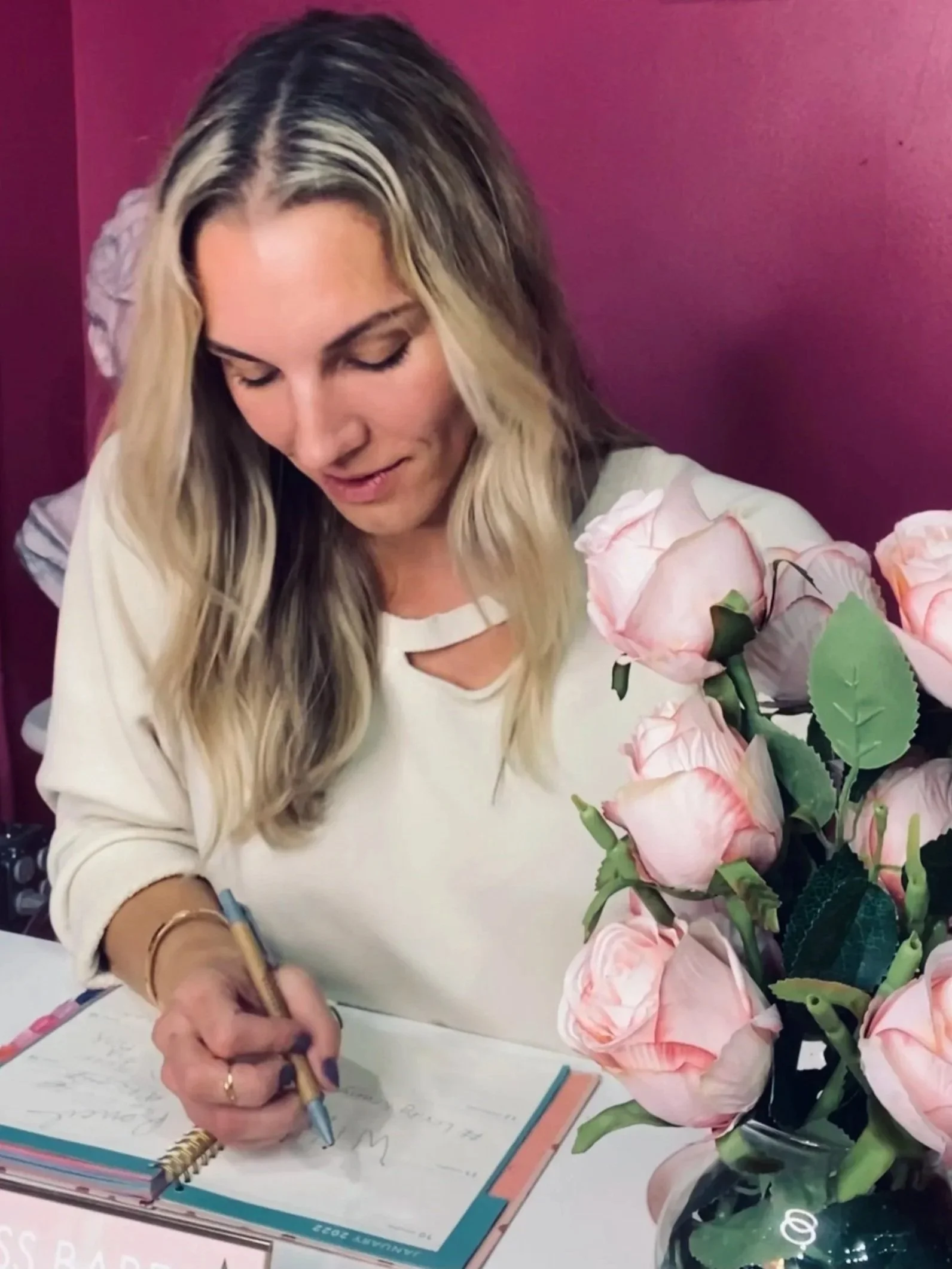 Woman with blonde hair signing a book or card at a table adorned with a bouquet of pink roses.
