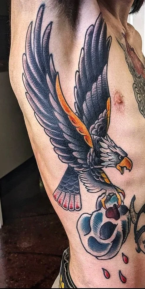 Tattoo of an eagle with wings spread, clutching a heart with blue and white shading, and red drops below.