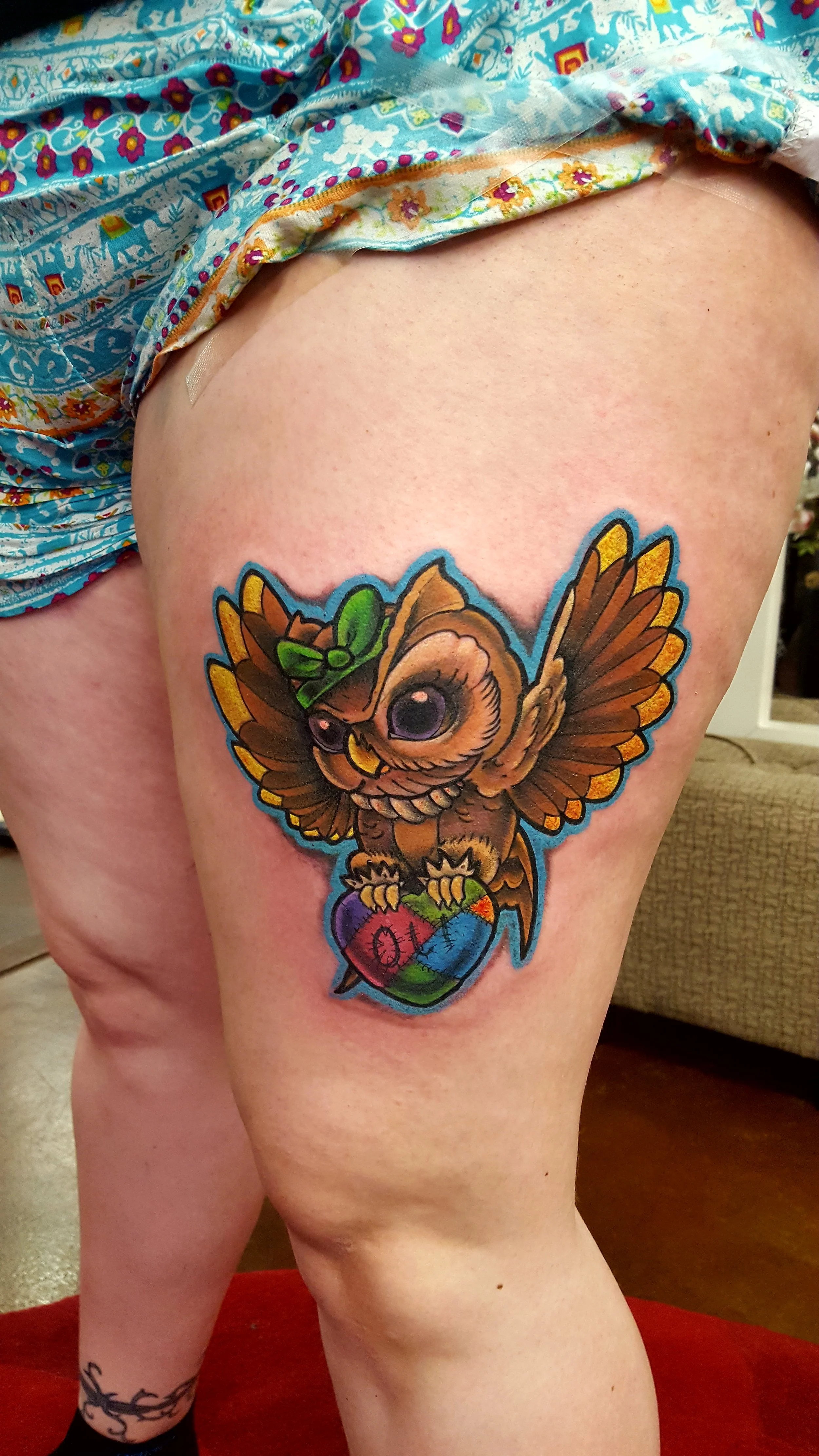 Colorful tattoo of an owl with large eyes, wearing a green bow, holding a wrapped heart-shaped object decorated with rainbow colors, on the person's upper thigh.