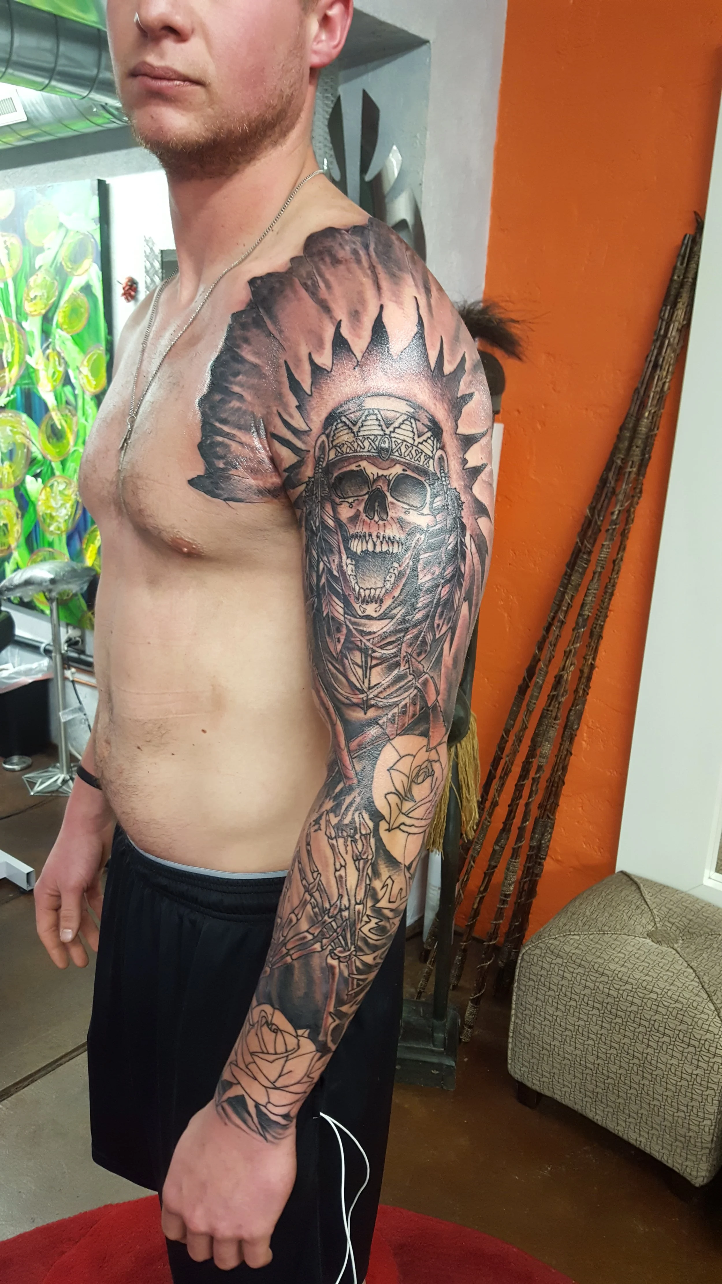 A shirtless man with a large detailed tattoo on his right arm and shoulder, featuring a skull with a feathered headdress and roses, standing indoors near an orange wall and a patterned chair.