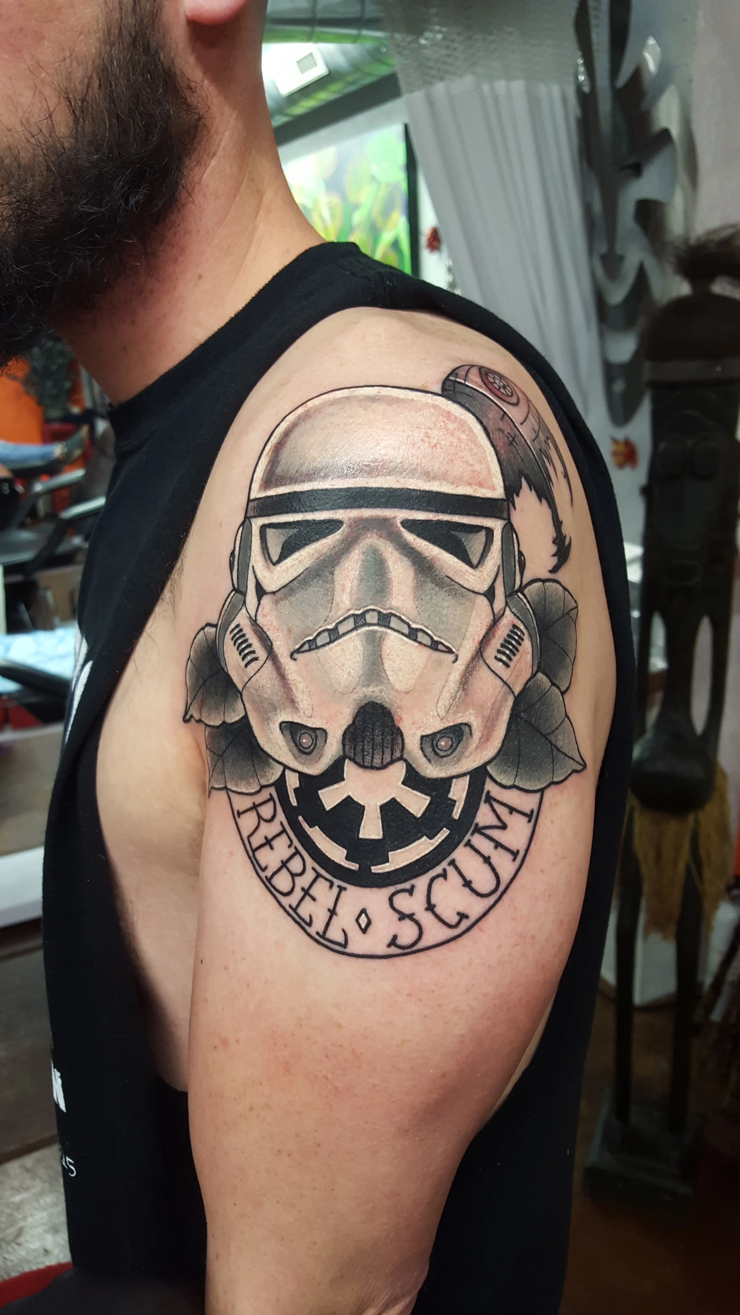 Tattoo of a Stormtrooper helmet from Star Wars with a skull design, leaves, and a circular banner reading 'Rebel Scum' on a man's shoulder.