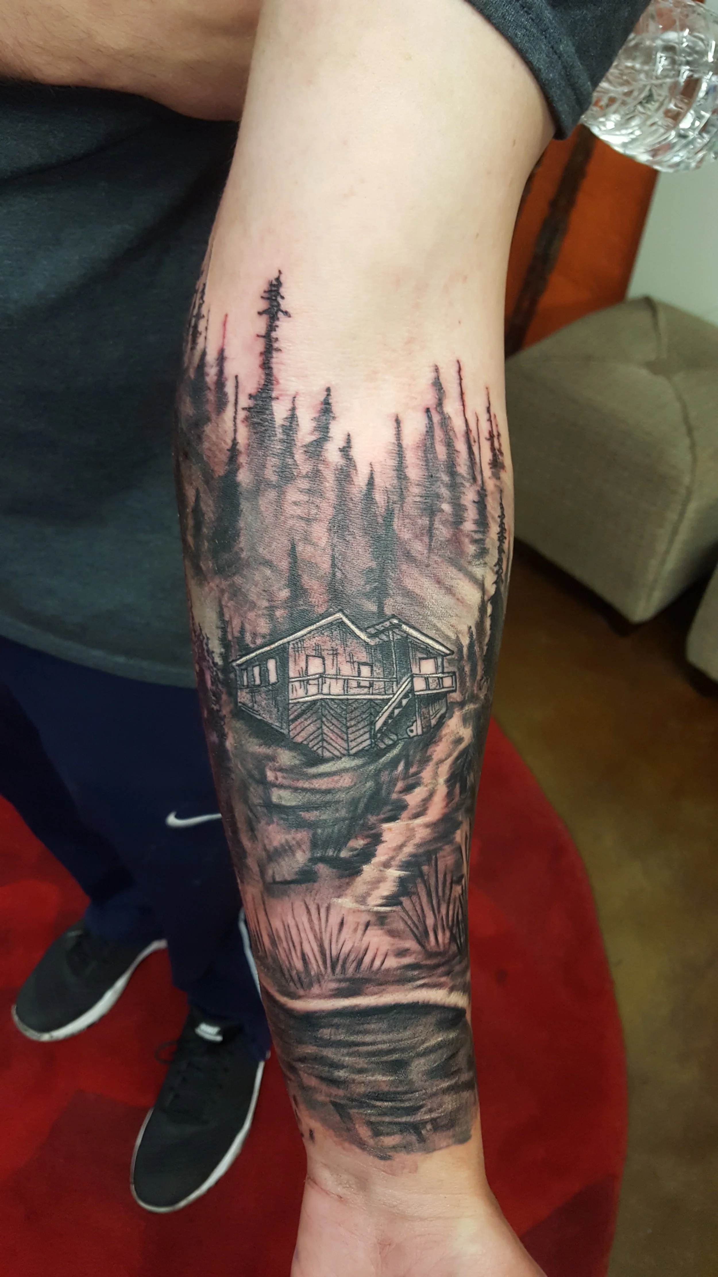 Black and gray tattoo of a cabin surrounded by trees and a mountain scene, with a body of water reflecting the landscape.