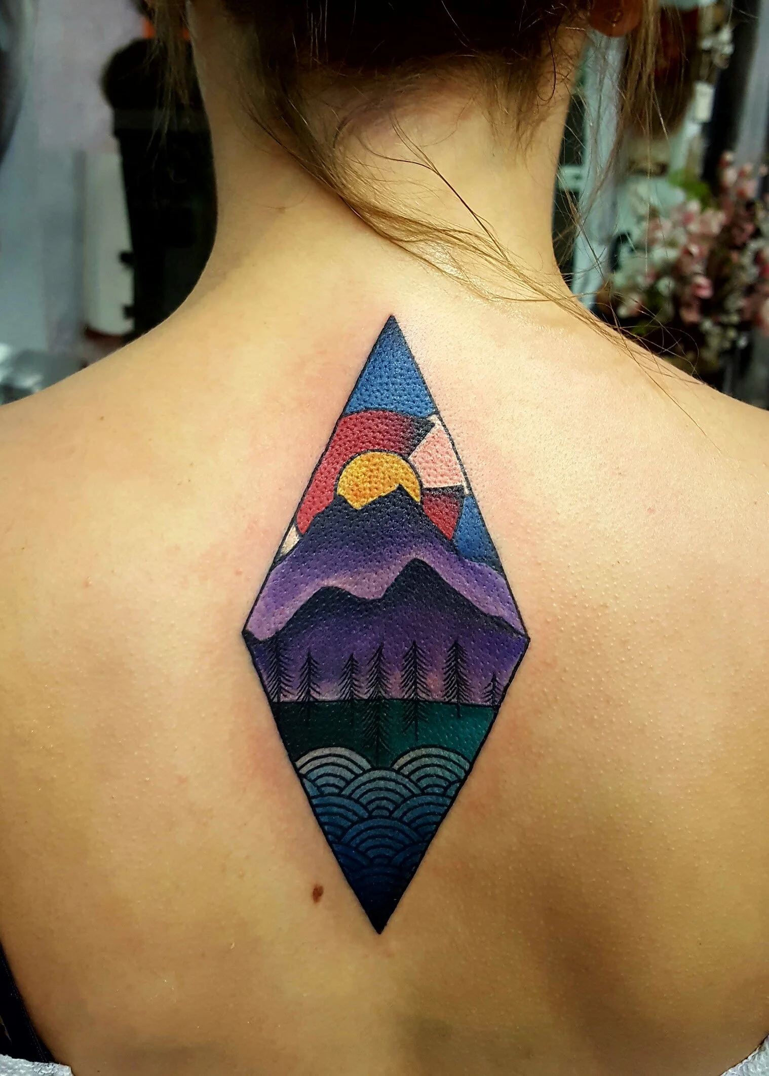 Tattoo on person's upper back featuring a stylized landscape with mountains, a sun, trees, and water, all within a diamond-shaped design.