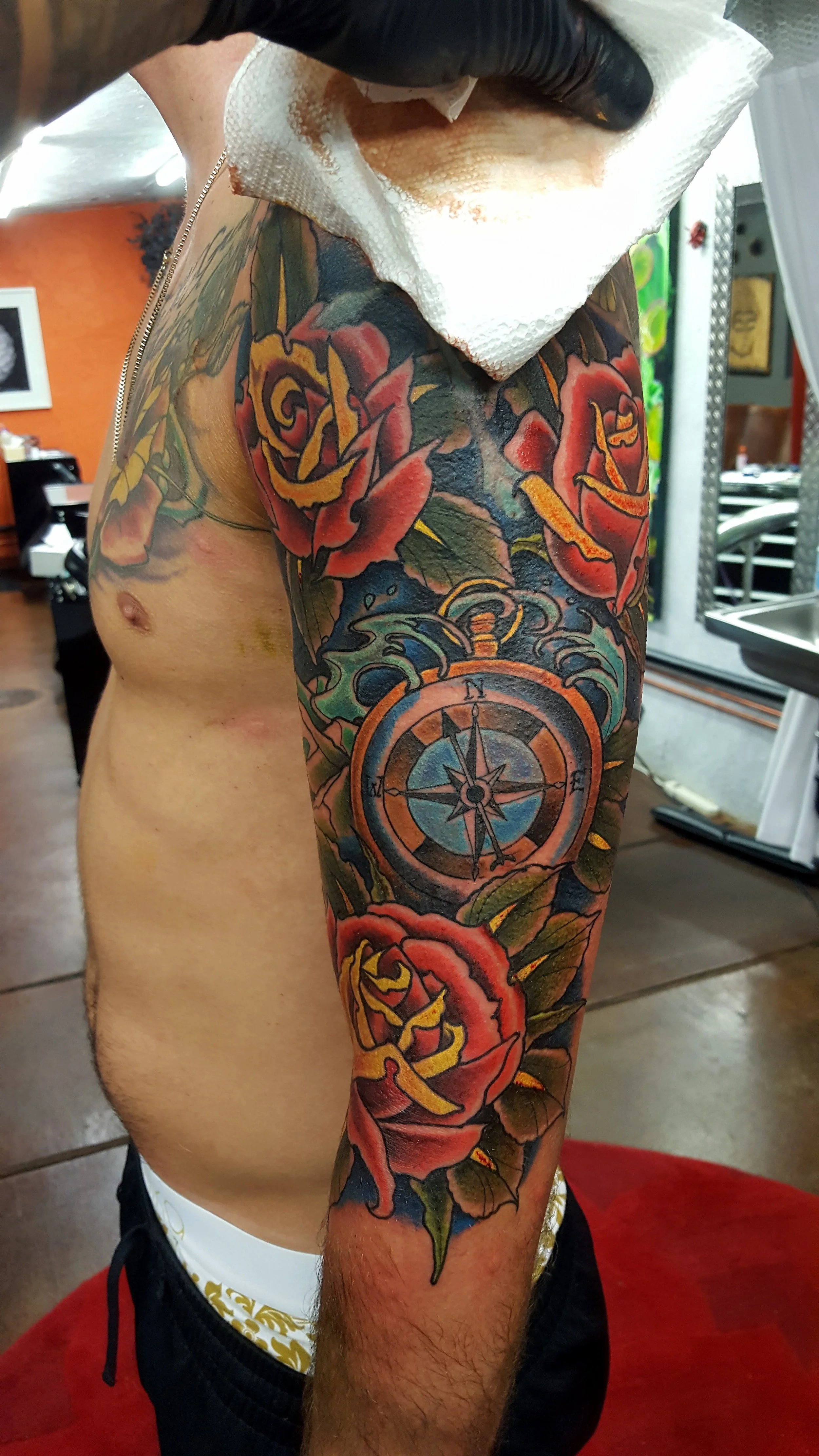 A person's upper arm with a colorful tattoo featuring roses, a compass, and swirling patterns, in a tattoo studio.