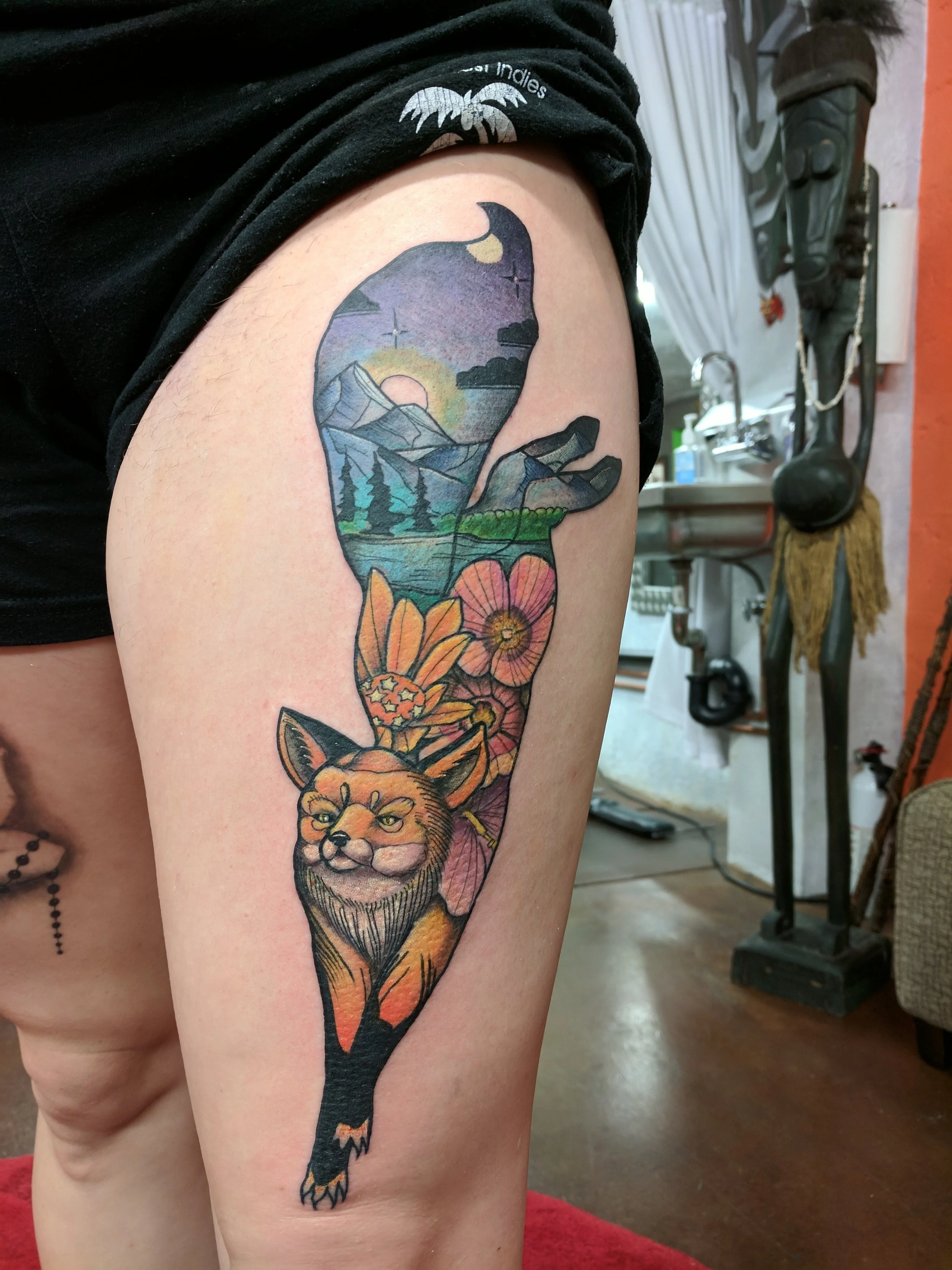 Tattoo of a fox with landscape and floral elements on person's upper arm. The landscape includes mountains, a sunset, trees, and clouds, while the floral elements consist of flowers and leaves.