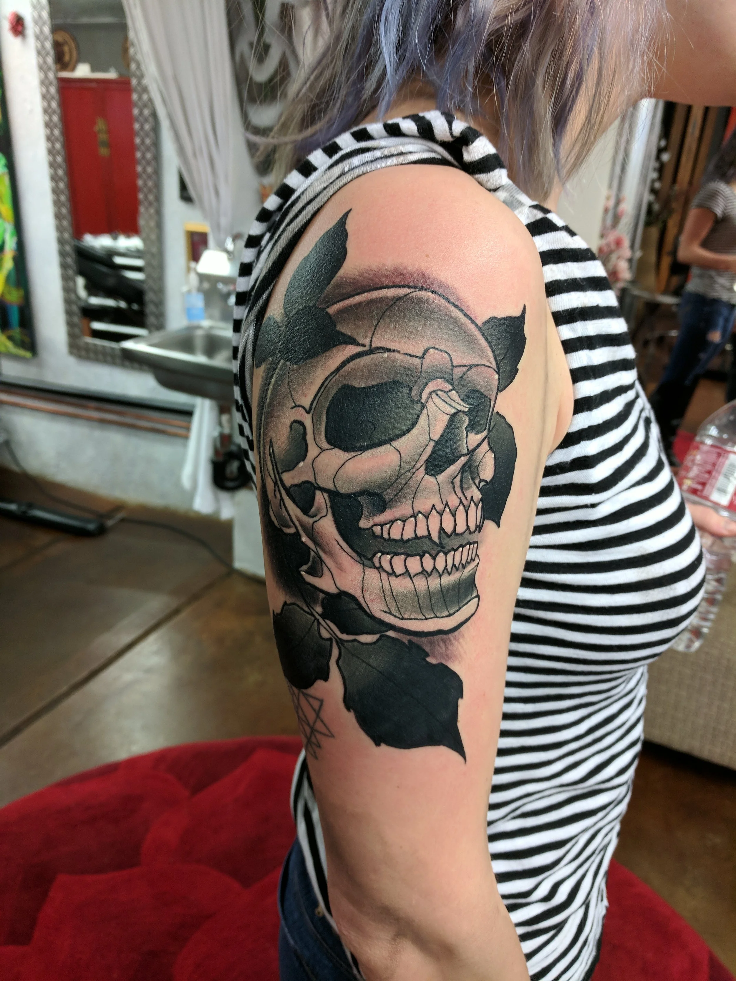 Black and gray tattoo of a skull with leaves on upper arm.