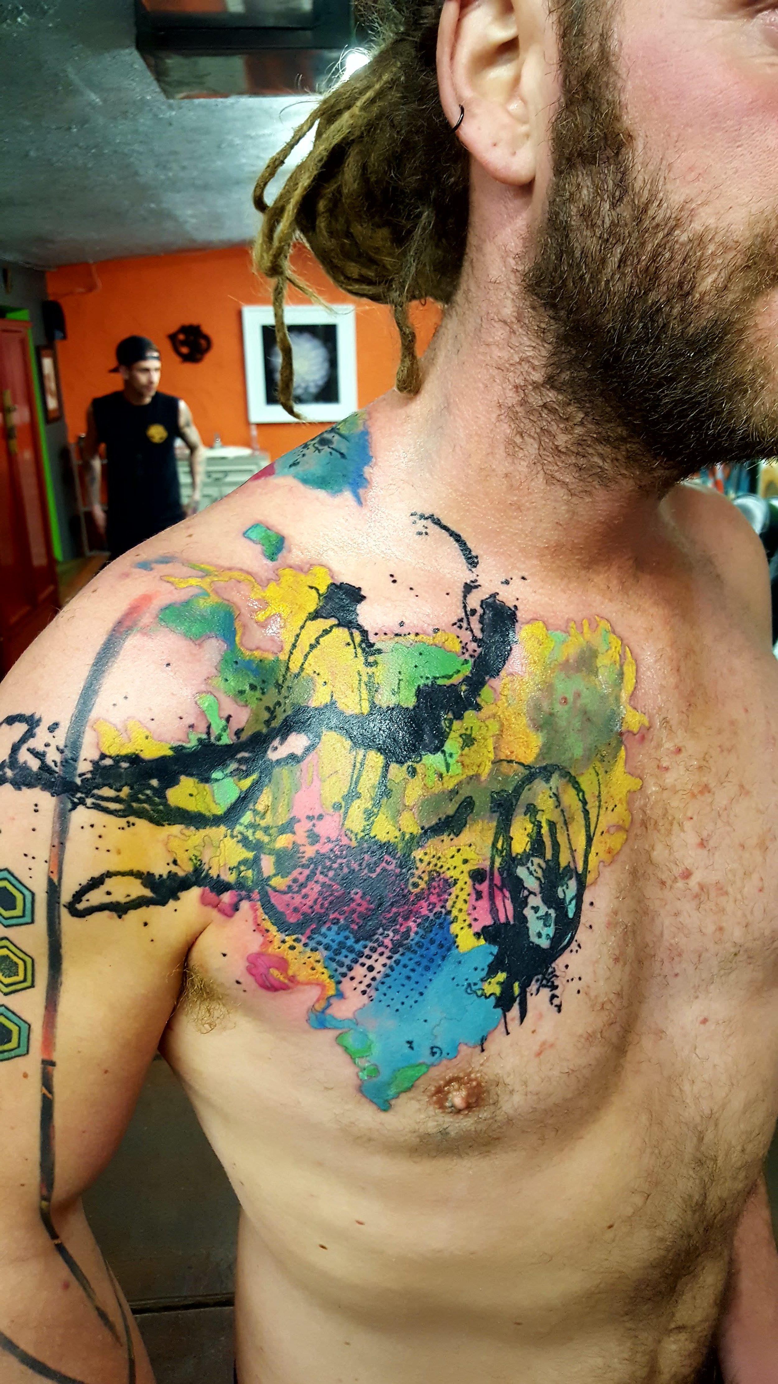 A shirtless man with colorful abstract tattoos on his shoulder and chest, featuring splashes of yellow, pink, blue, and green ink, with black ink outlines. In the background, part of an orange wall, a framed black and white artwork, and a man wearing