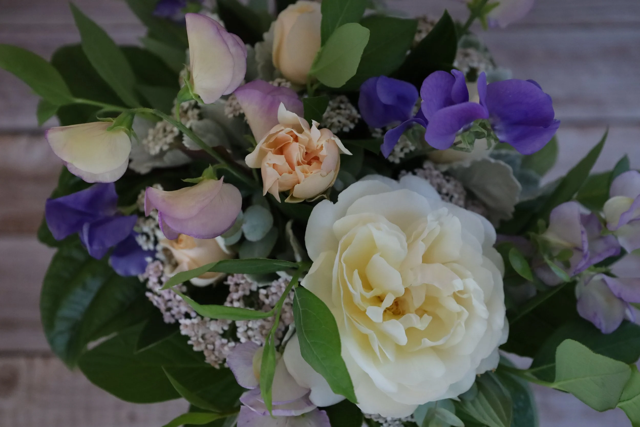 A colorful bouquet of various flowers against a gray background, including dahlias, coneflowers, and other blooms, accented with green leaves and small berries.