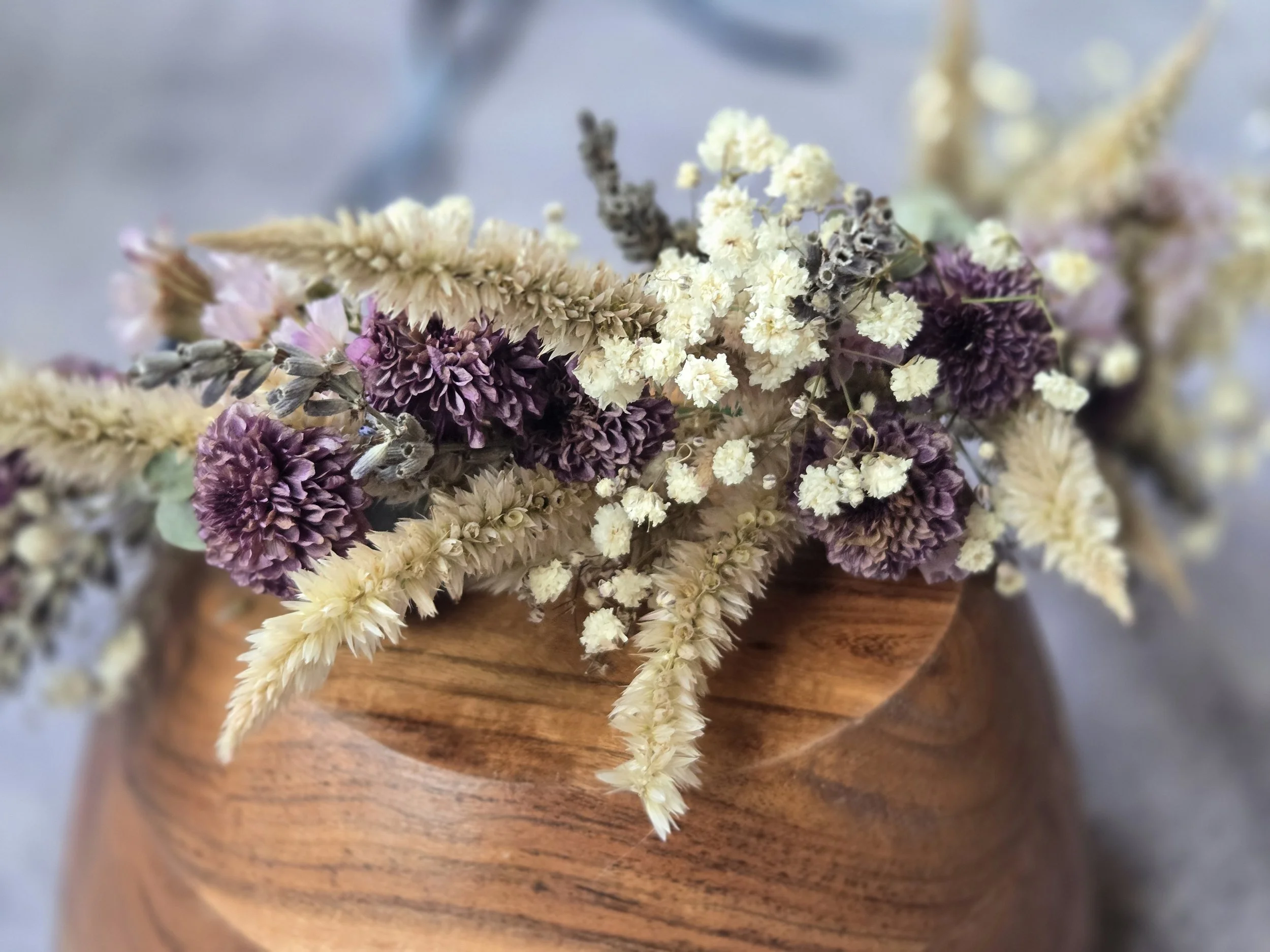 A floral arrangement with various colorful flowers in a glass vase, set on a white tablecloth. On the table, there's a metal container labeled 'Flowers & Garden' holding scissors and twine, along with a sign that reads 'Flower Bar make a bouquet to take on your way!' with colorful flower drawings. In the background, people are sitting on a deck, and some are visible from the shoulders up.