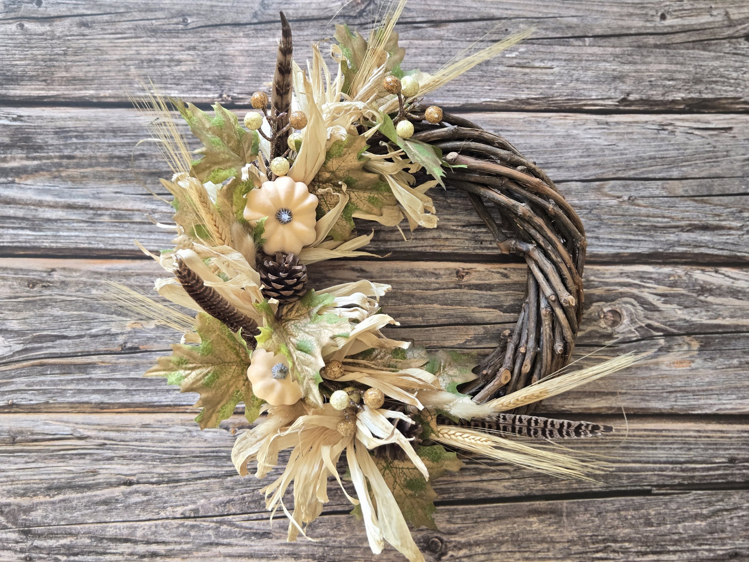A decorative autumn wreath made of intertwined brown twigs, adorned with beige tree bark flower, maple leaves, pine cone, feathers, and small golden embellishments, placed on a weathered wooden surface.