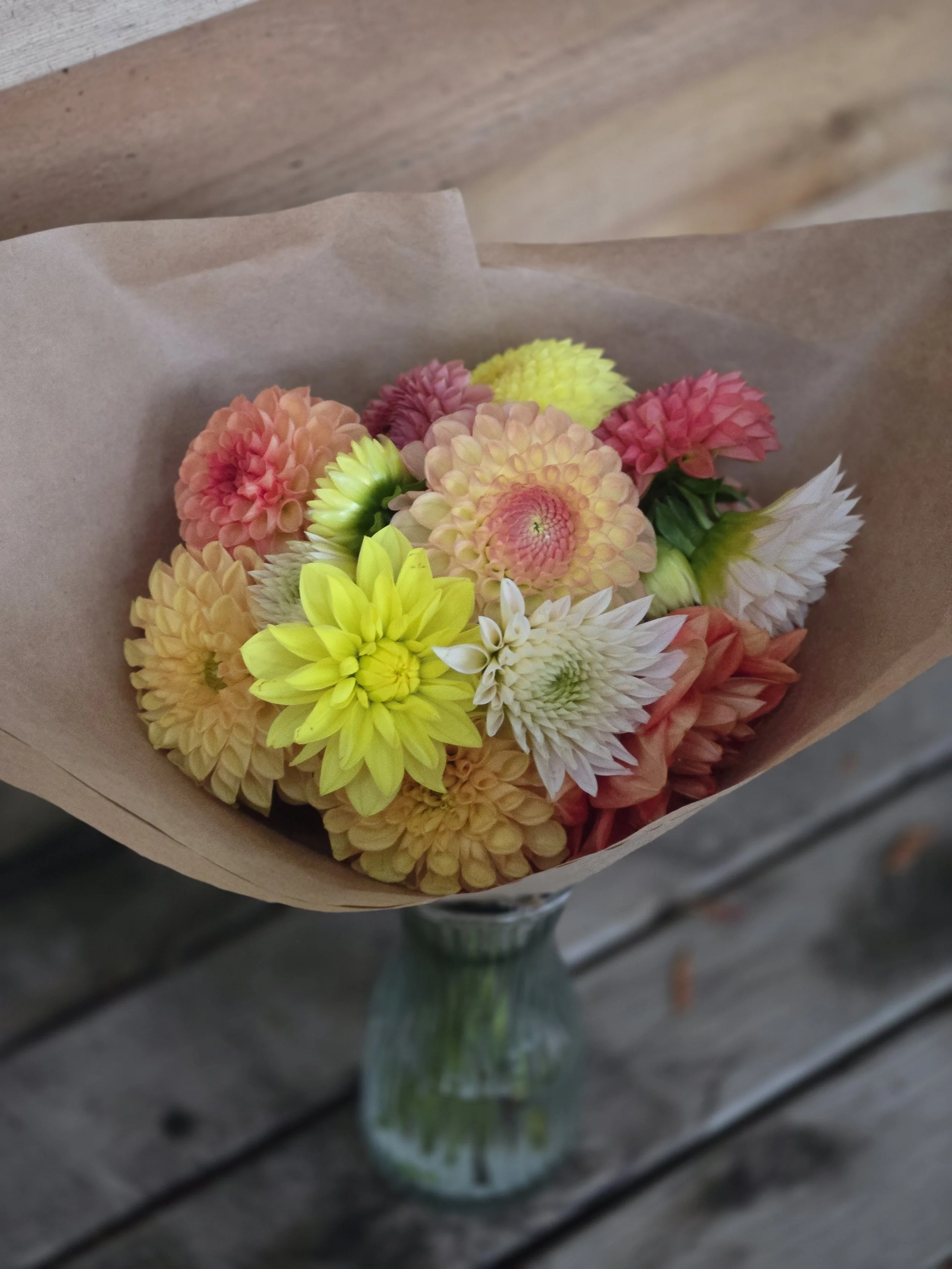 Colorful bouquet of mixed flowers in a glass vase, wrapped in brown paper, placed on a wooden surface.