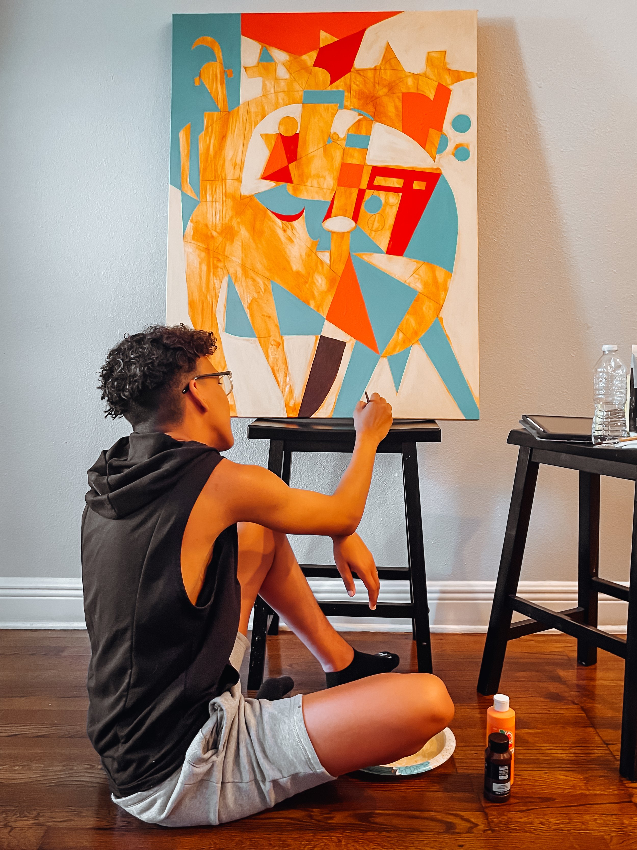 Young person sitting on the floor painting a large abstract canvas with bright orange, blue, red, and cream geometric shapes.