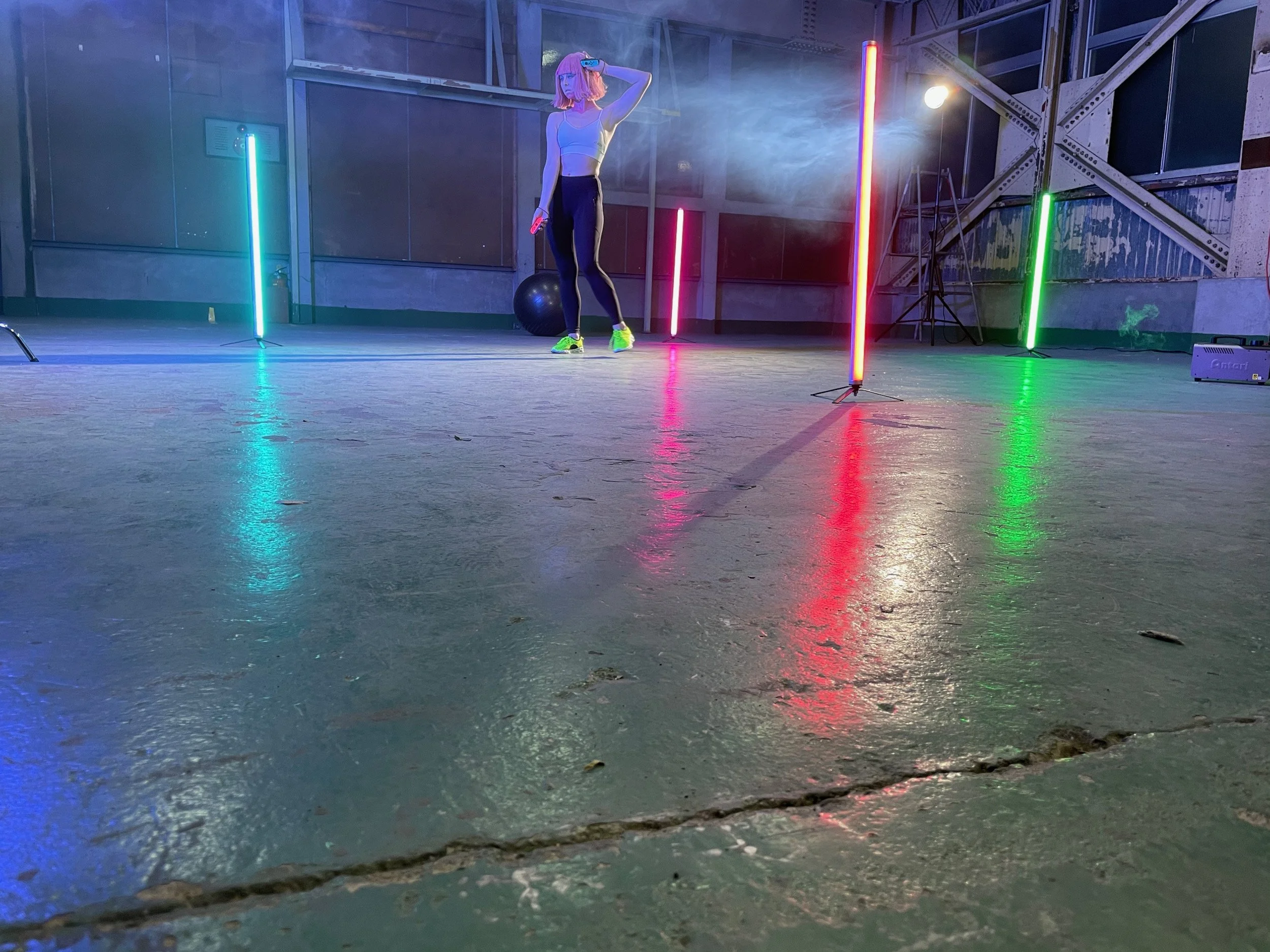 A woman with pink hair stands in a dance studio with neon light bars in various colors, multiple smoke effects, a black exercise ball, and industrial-style exposed beams.