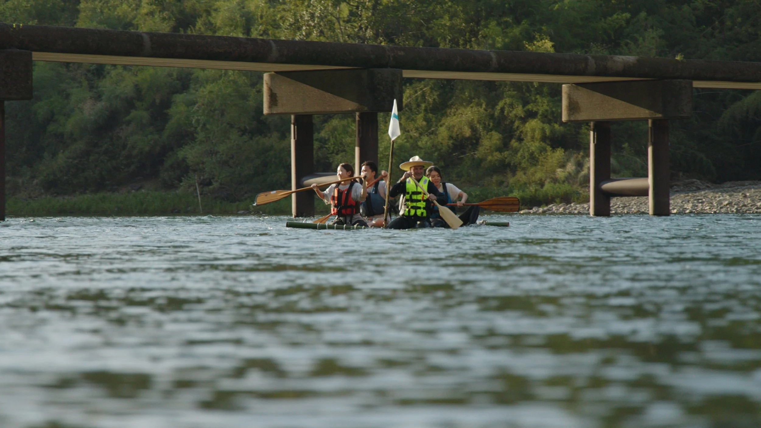 A group of five people kayaking on a river under a bridge, with lush green trees on the riverbank in the background.