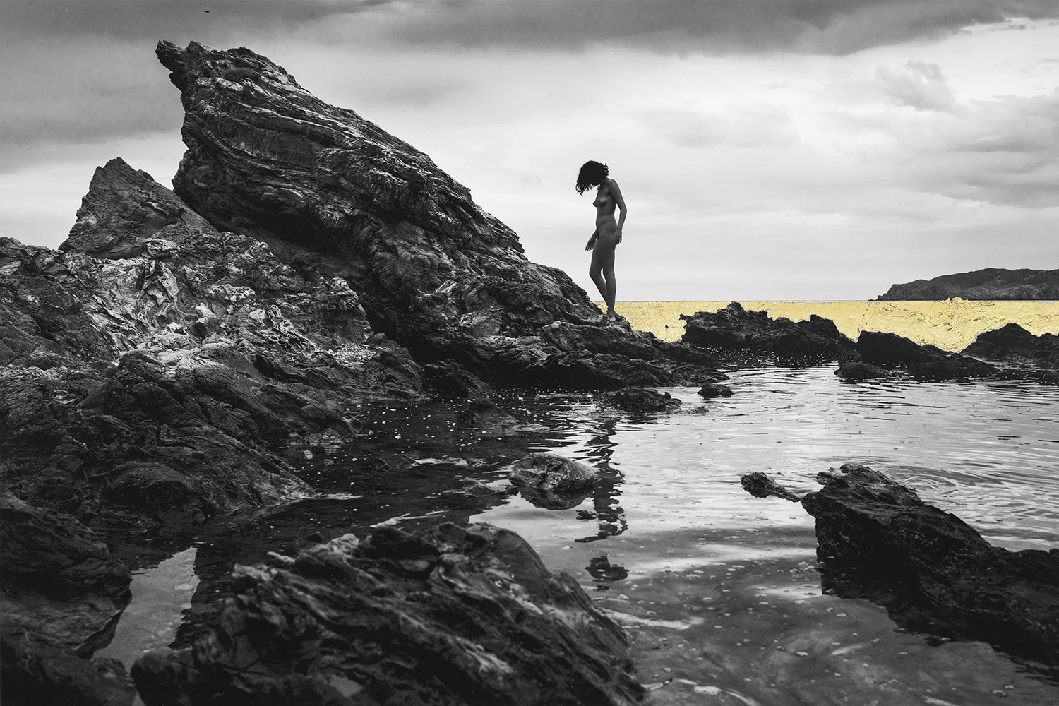A black-and-white photo of a woman standing on rocky shoreline by the water, with a large jagged rock formation nearby and a cloudy sky overhead. A strip of color highlights the distant beach or background.
