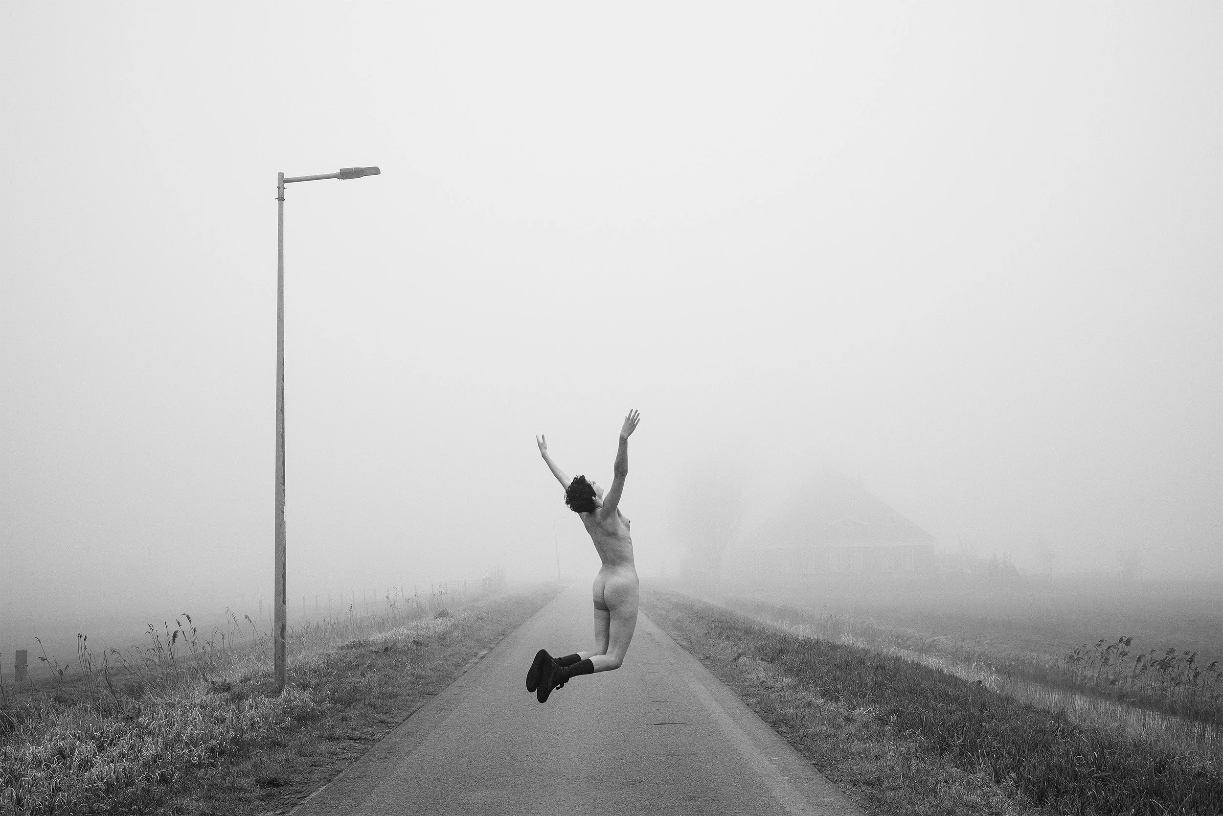 A person with curly hair jumpin on a road in an outdoor setting, reaching their arms up towards another person who is floating upside down in front of them, with grass on either side of the road and a streetlamp in the background, in black and white.