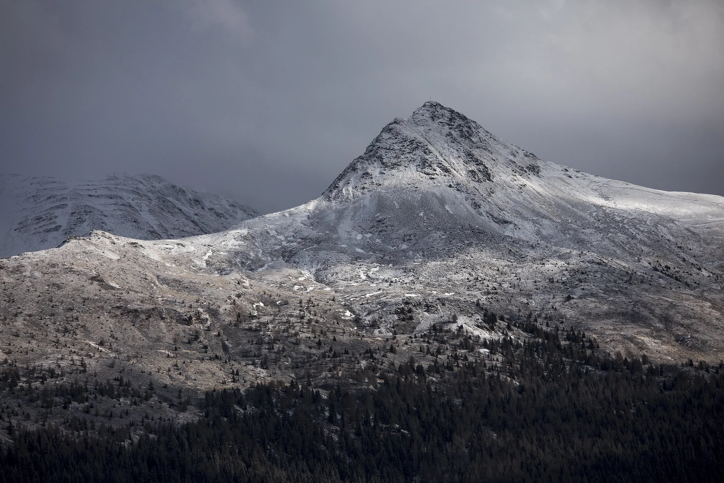 Snow-covered mountain with dark forest at the base and a cloudy sky.