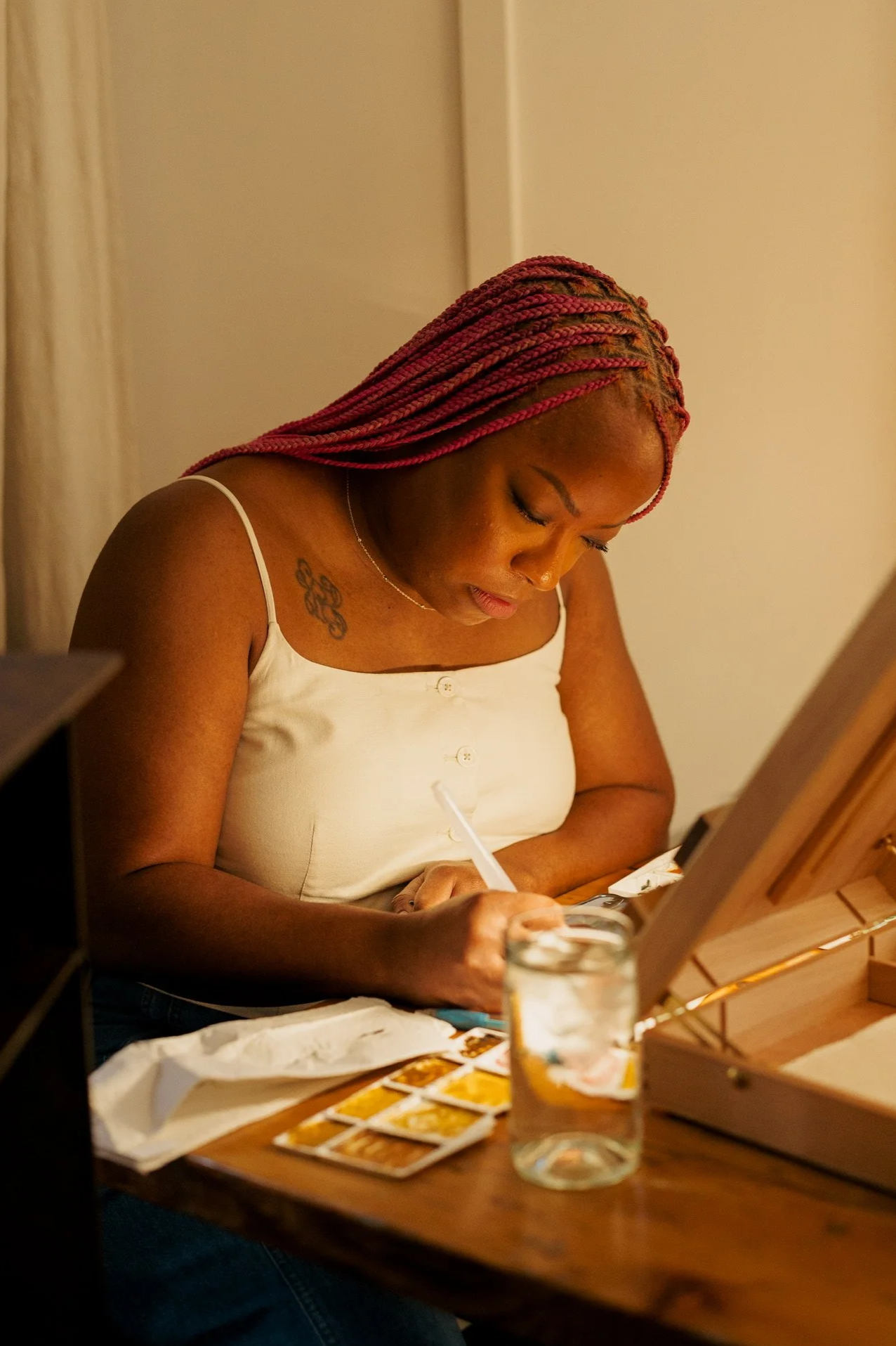 A woman with pink braided hair is painting abstract portraits at a table with watercolor paints, a glass jar of water, and a wooden painting easel.
