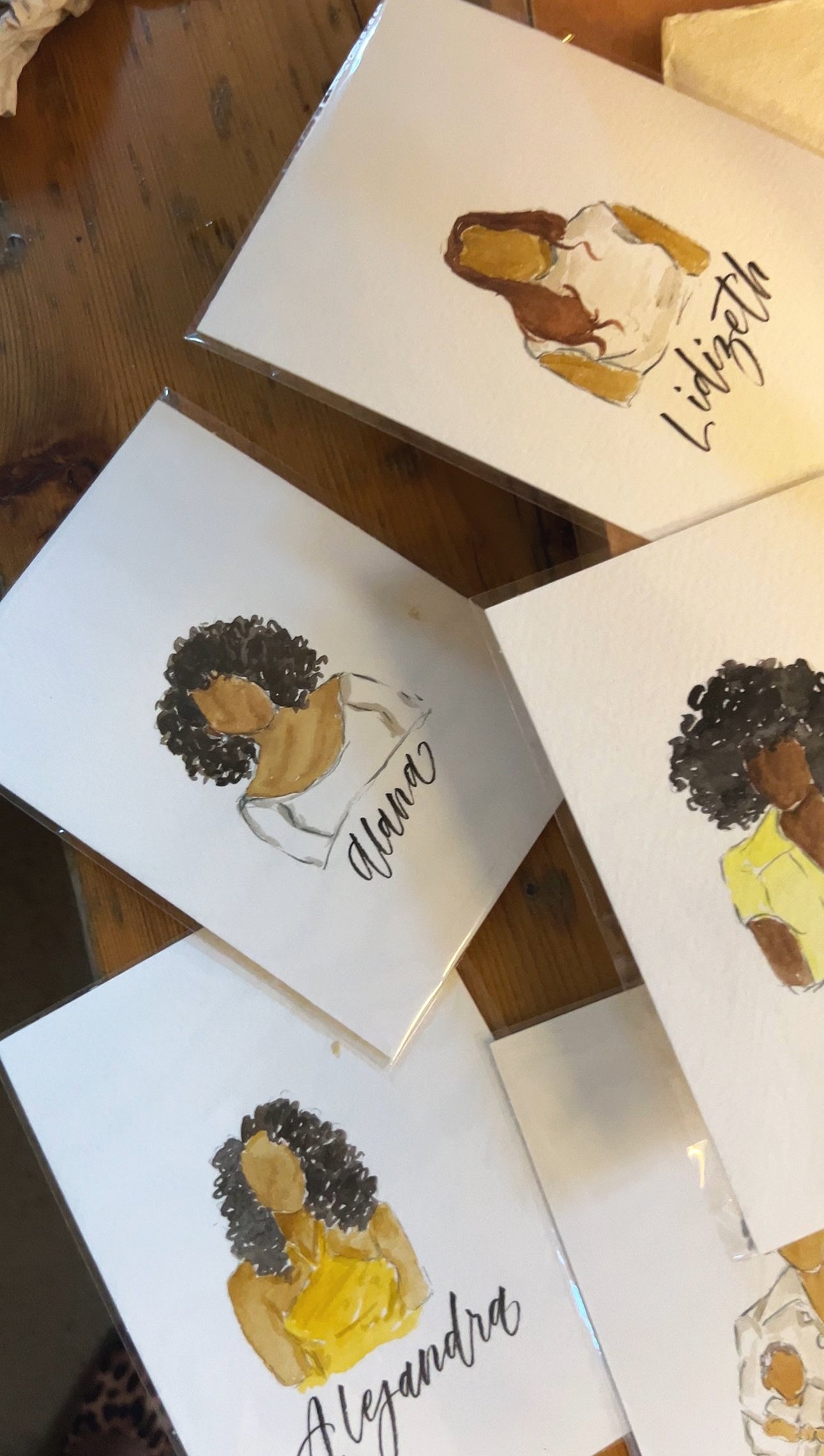 Watercolor illustrations of women with personalized names on cards, displayed on a wooden surface.