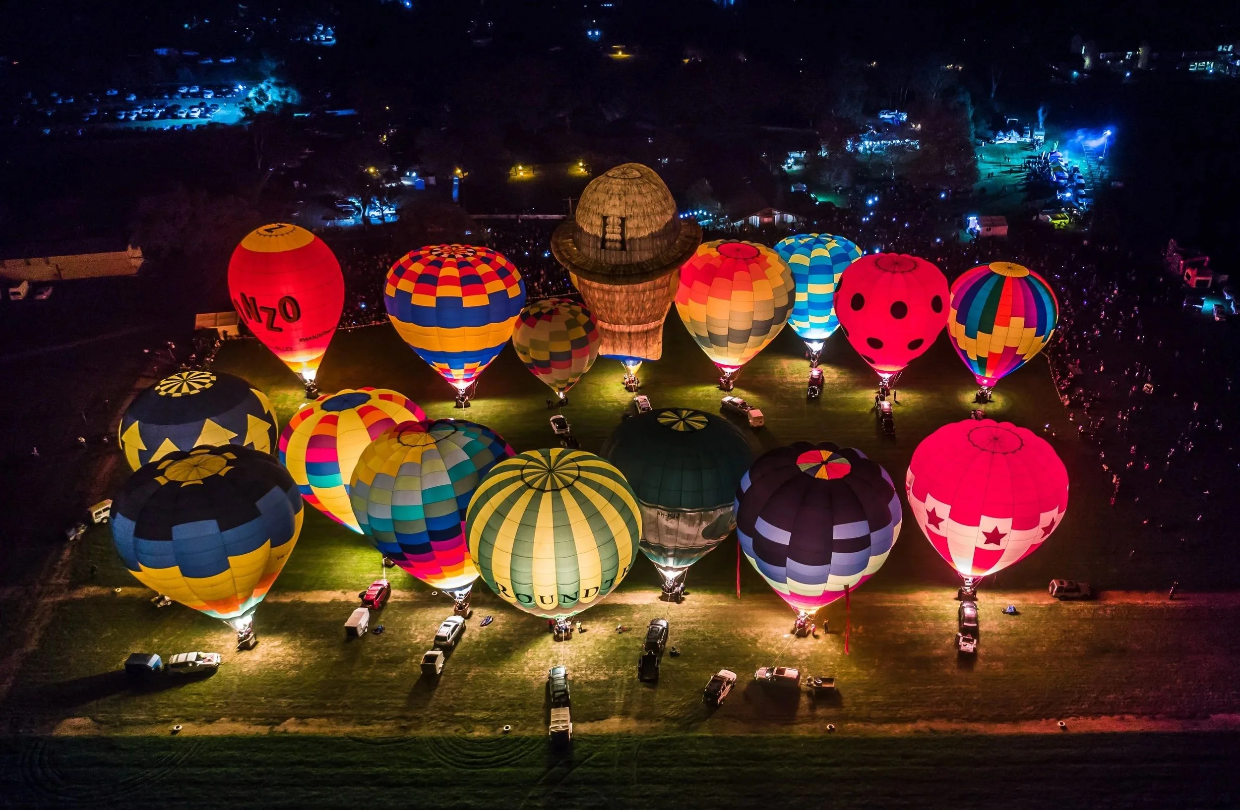 Hot air balloons illuminated at night on a field, including a hot air balloon shaped like a hat and one with ladybug and star patterns, with a crowd and vehicles surrounding the area.