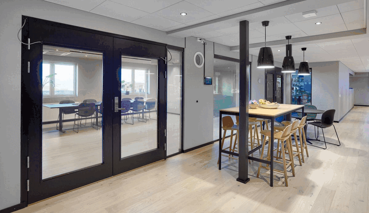 Interiør av et moderne kontor eller møtelokale med et høyt spisebord og stoler, og et rom med møteromsdører med glassvinduer.