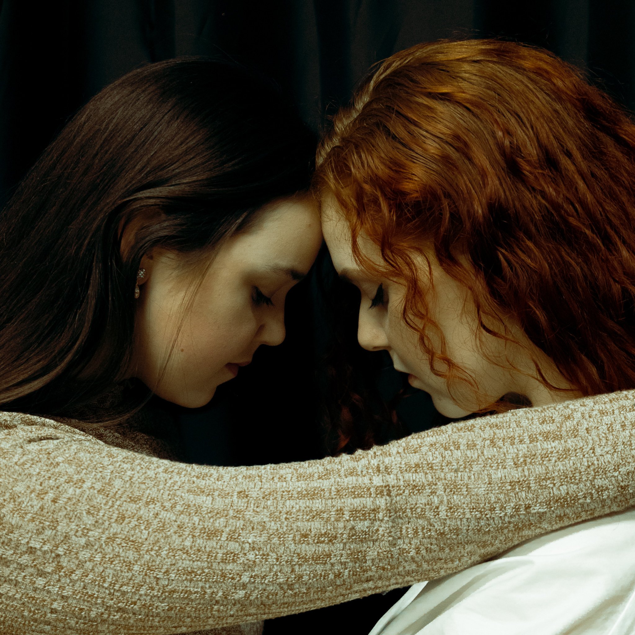 Two women with closed eyes and foreheads touching, resting their heads on each other's arms.