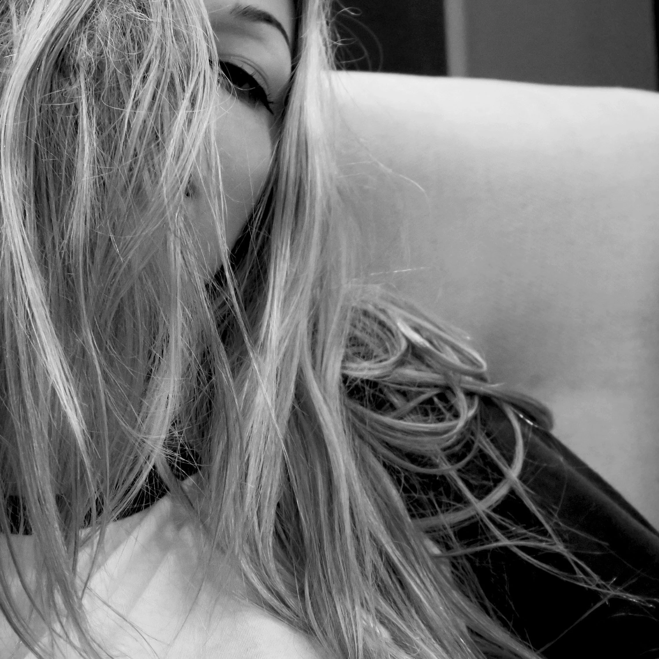 Close-up black and white photo of a woman with long, messy hair partially covering her face, leaning on a surface, with another person's shoulder visible in the background.