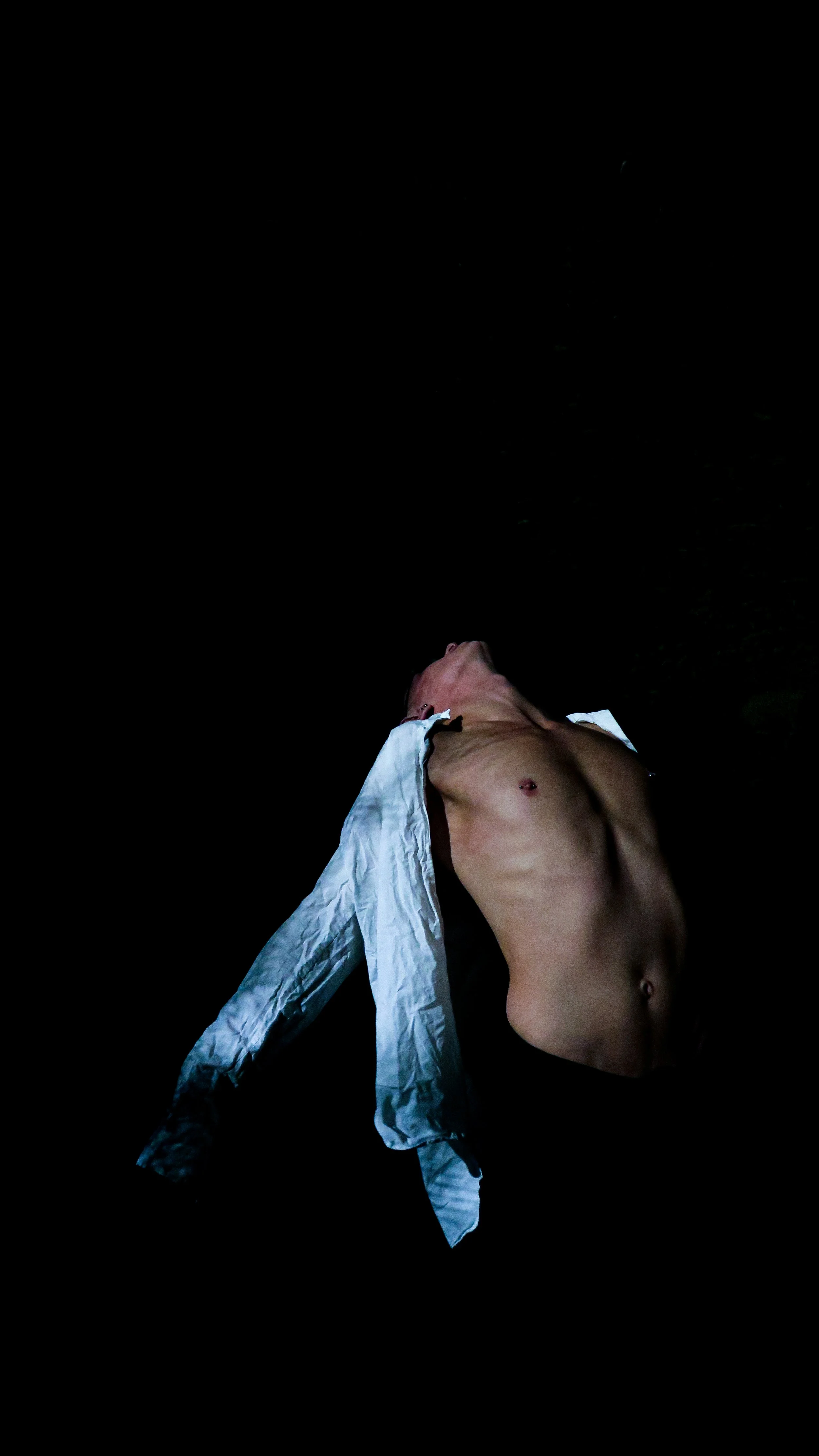 A shirtless person lying on their back with arms out and head tilted back against a dark background, wearing a white open shirt.