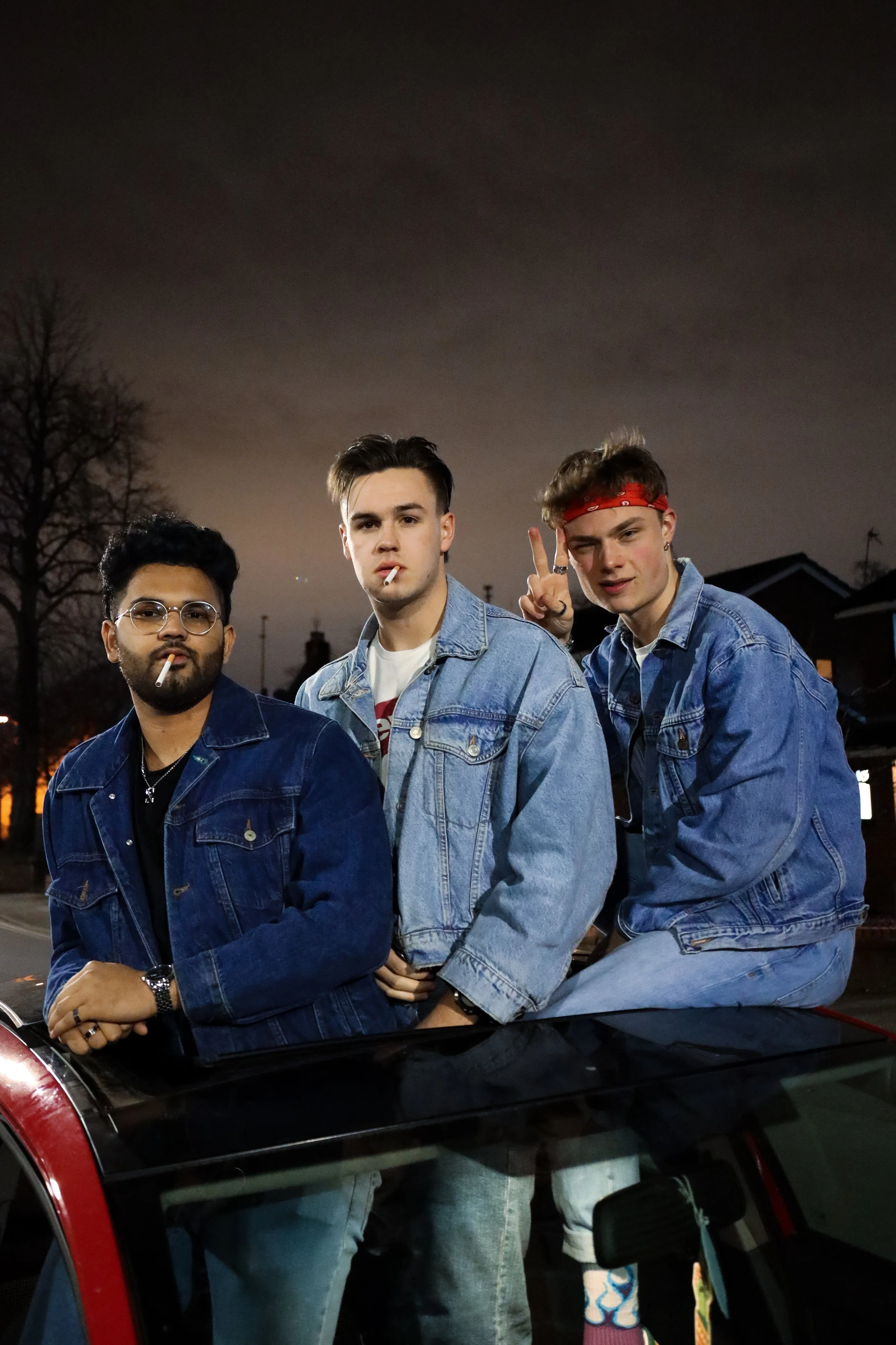 Three young men with cigarettes posing on a car at night, two of them making peace signs and wearing denim jackets.