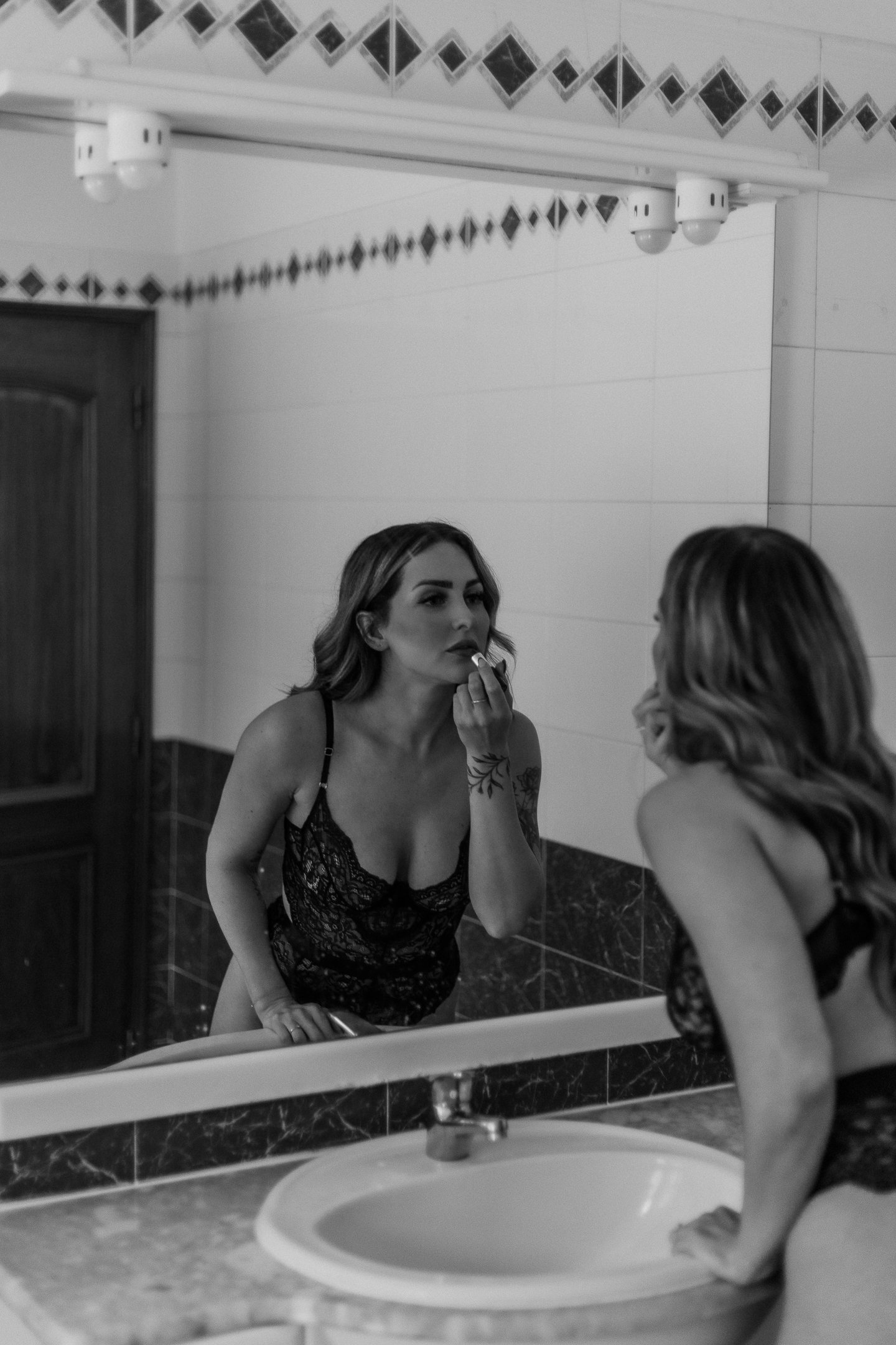 A woman in black lace lingerie looking into a bathroom mirror while applying lipstick, her reflection visible in the mirror.