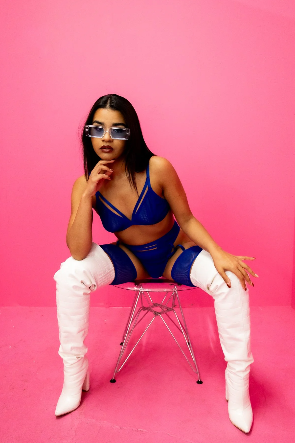 A woman wearing a blue lingerie set and white thigh-high boots, sitting on a clear acrylic chair against a pink background, with her hand on her chin and wearing sunglasses.