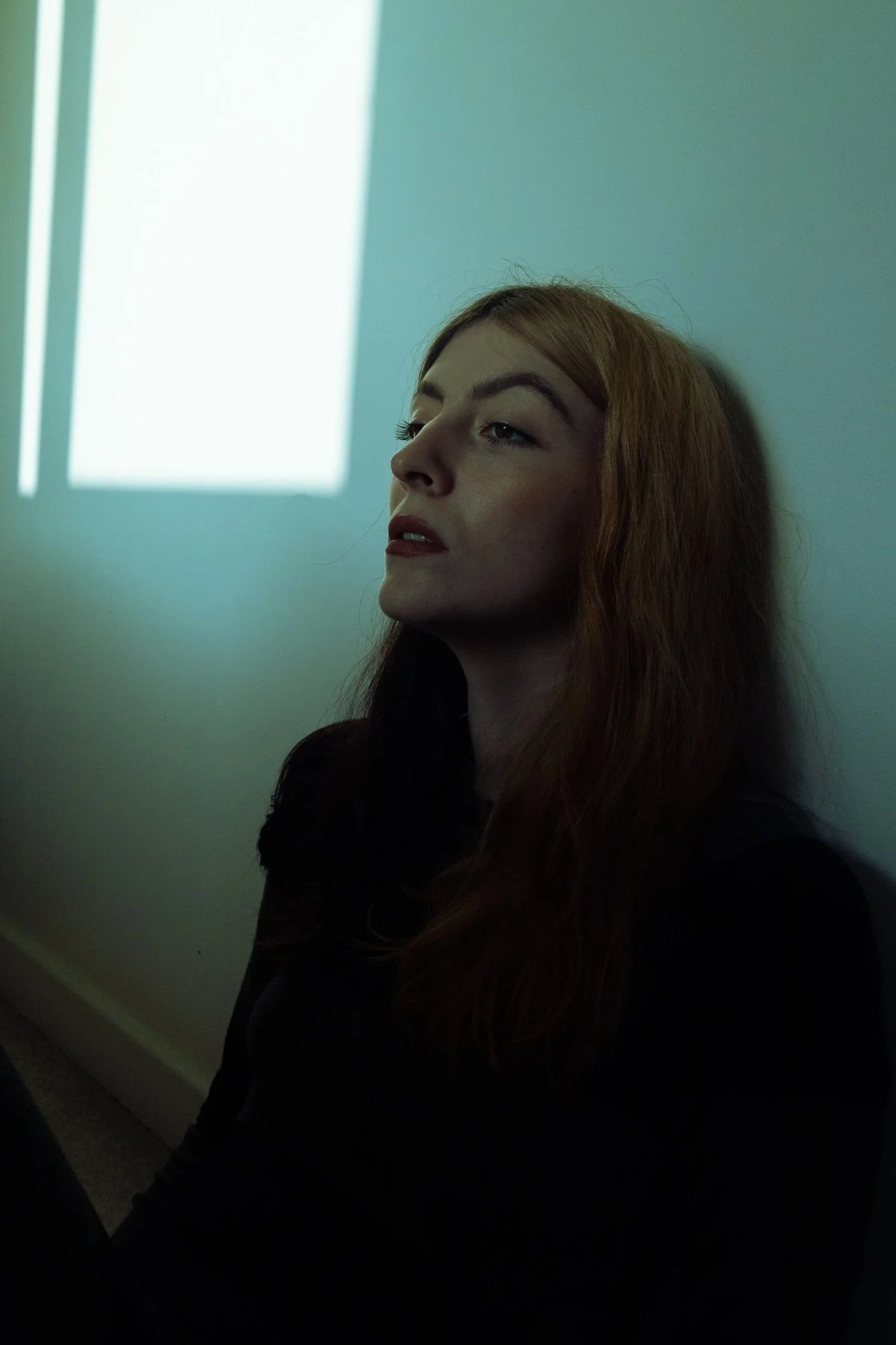 A young woman with red hair and light skin sits against a wall in a dimly lit room with sunlight streaming through a window, casting rectangular light patches on the wall behind her.