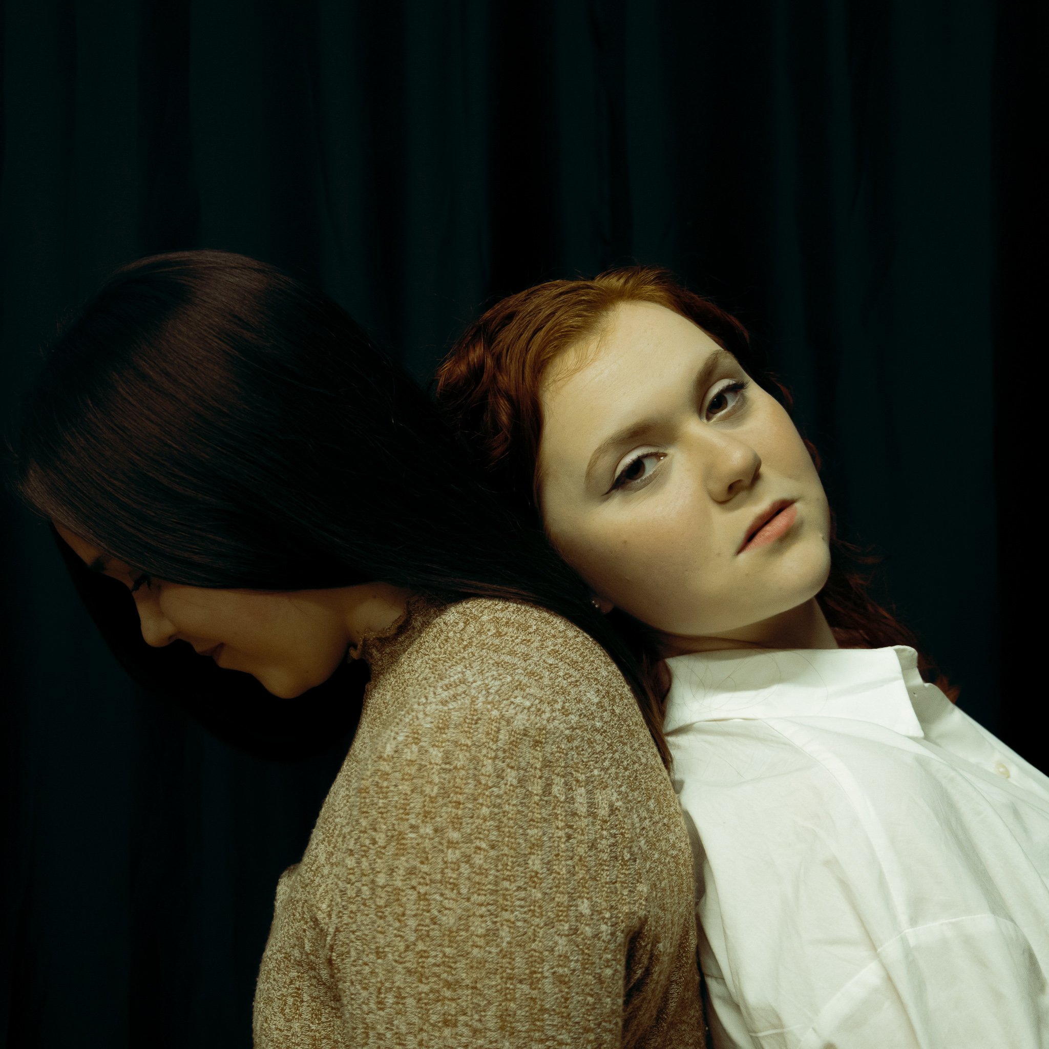 Two women sitting back to back against each other's shoulders, one with dark hair looking down and the other with red hair looking at the camera, in front of a dark background.