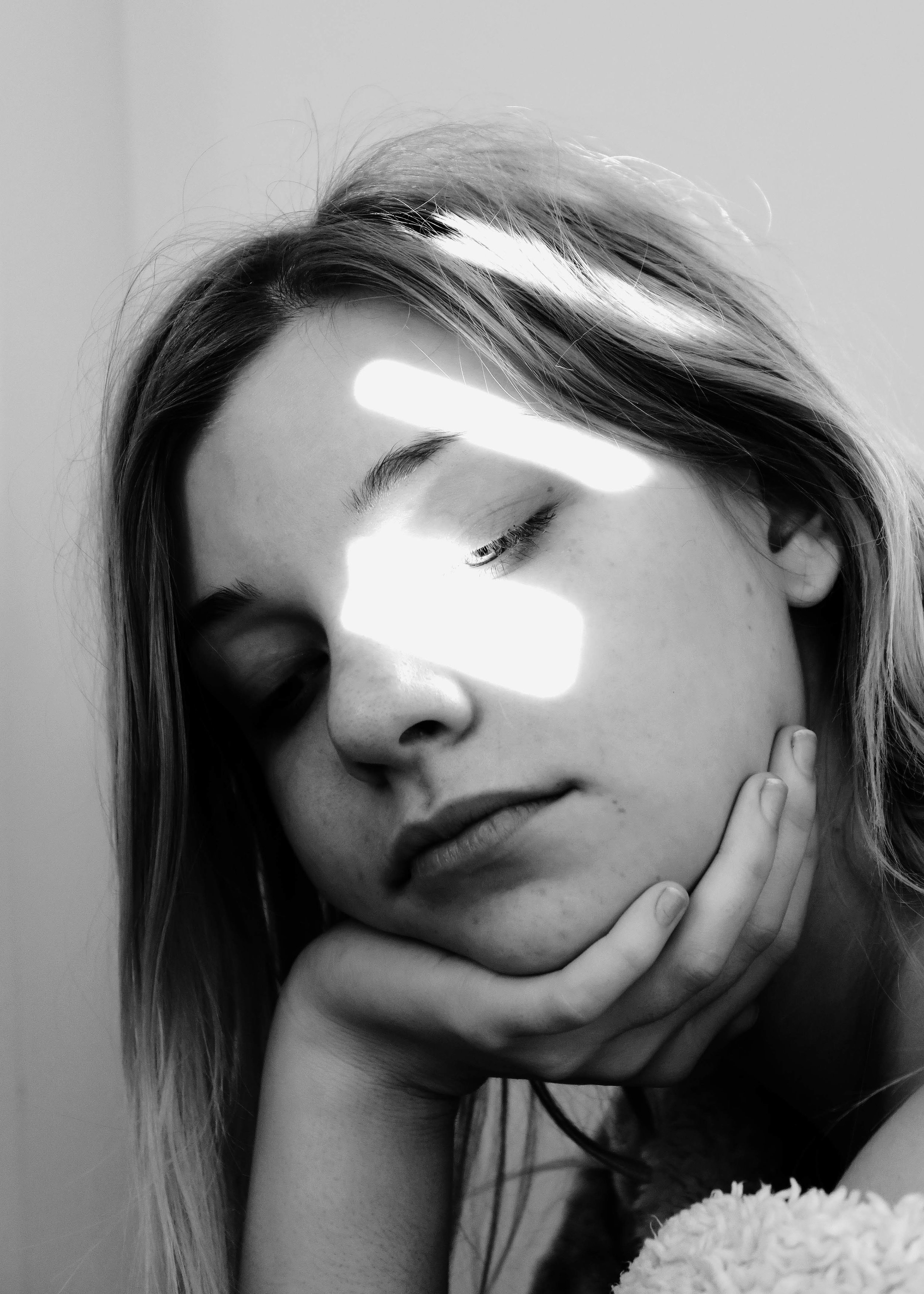Black and white portrait of a young woman with closed eyes, resting her chin on her hand, with light streaks crossing her face.