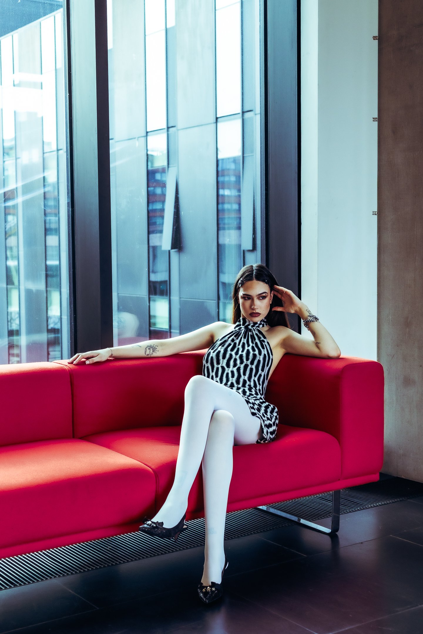 Woman sitting on a red sofa in a modern building with large glass windows, wearing a black and white patterned dress and white tights.