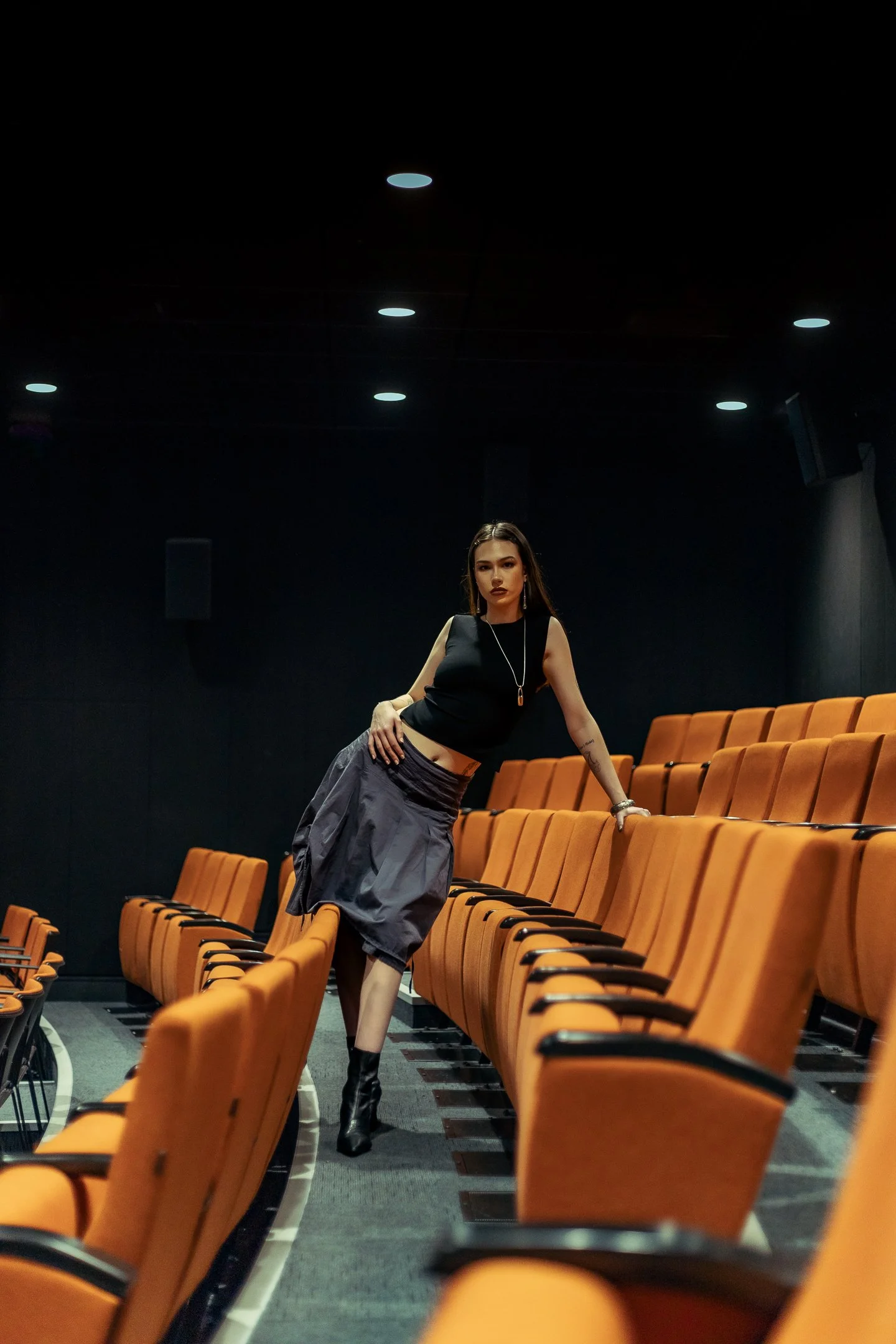 A woman in a black sleeveless top and gray skirt standing in an empty theater with orange seats.