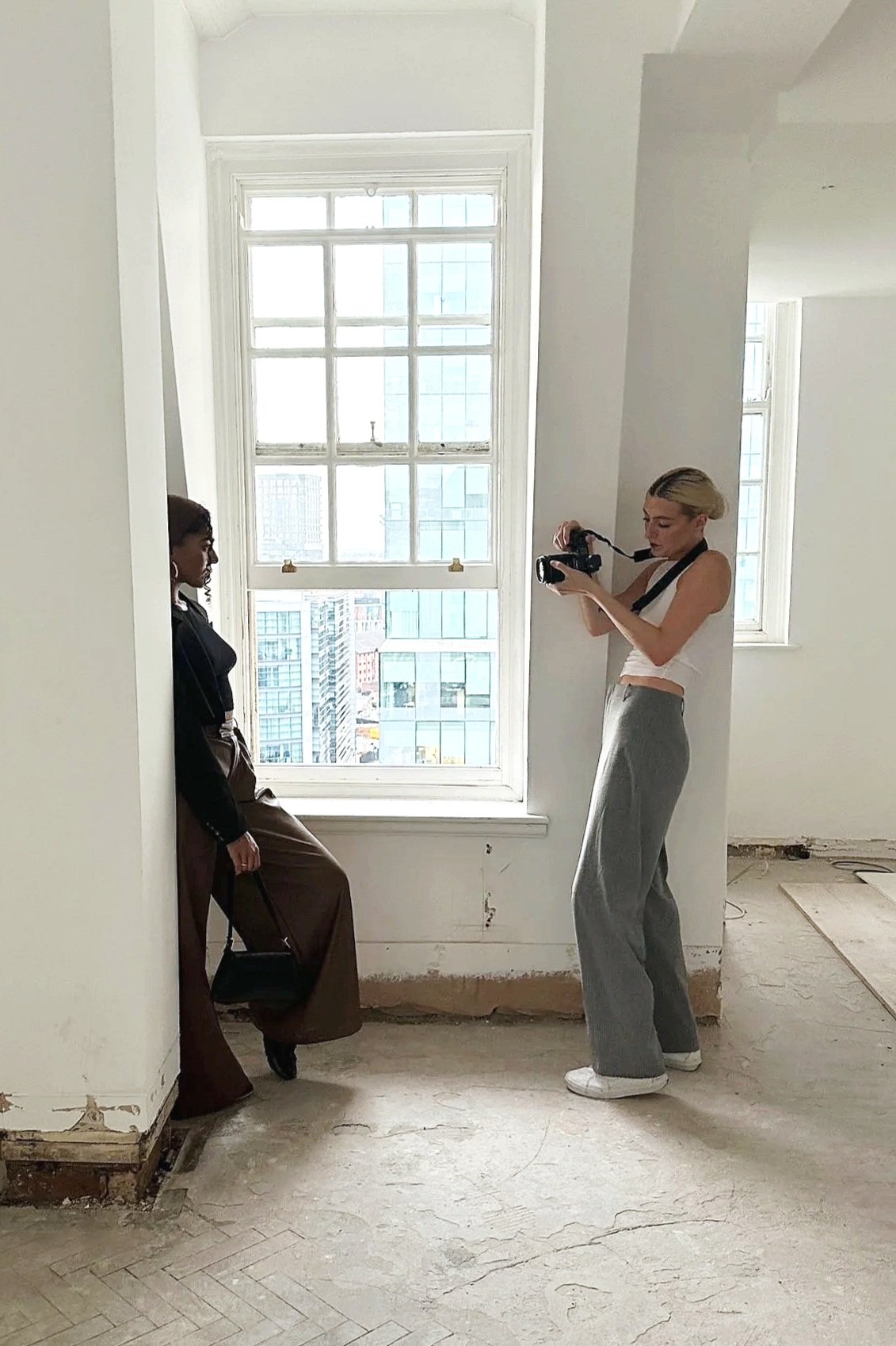 A woman with blonde hair taking a photo of a sitting woman with dark skin and curly hair against a large window with a city view in a minimally decorated room.