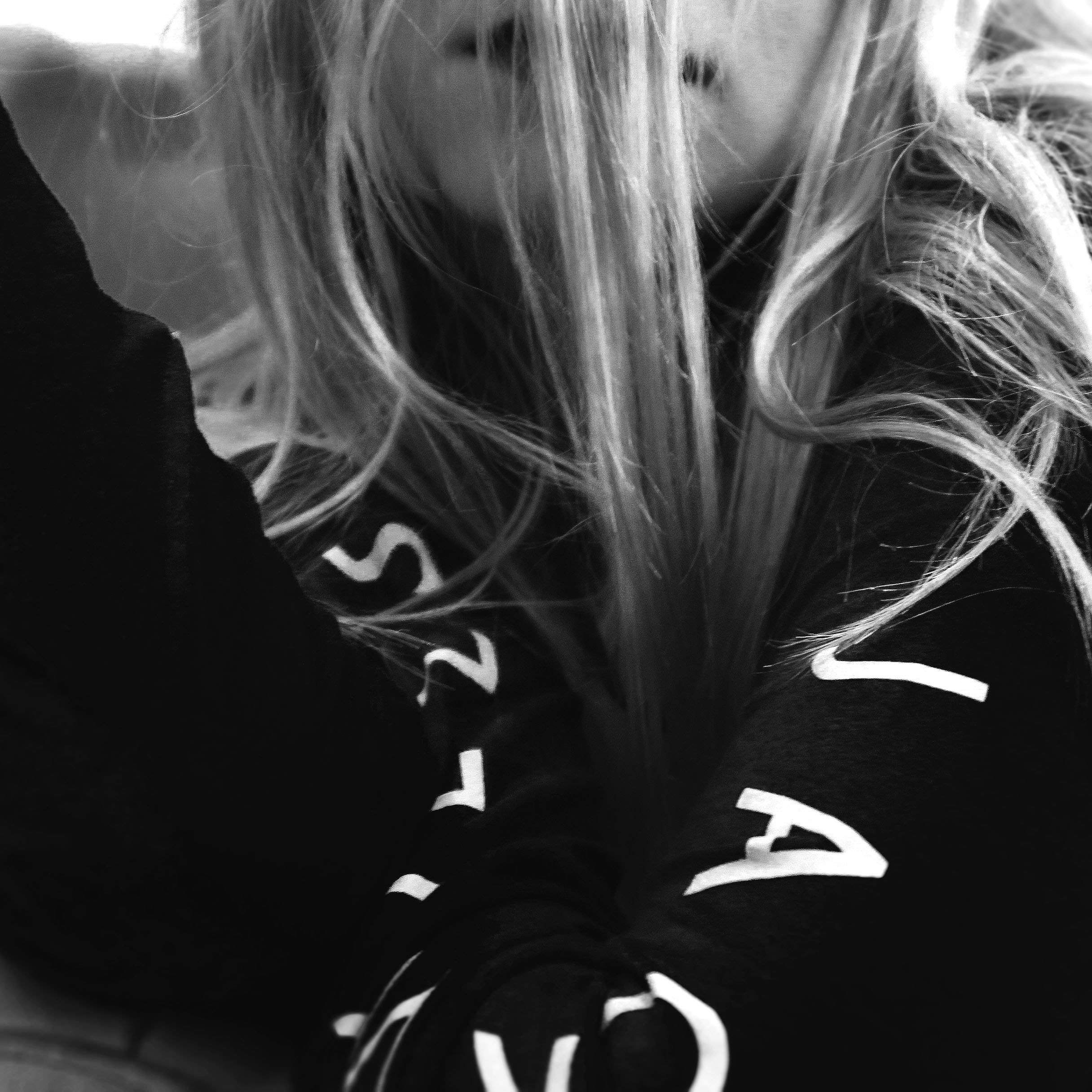 Close-up of a person's face partially obscured by long, wavy hair, wearing a dark hoodie with white text.