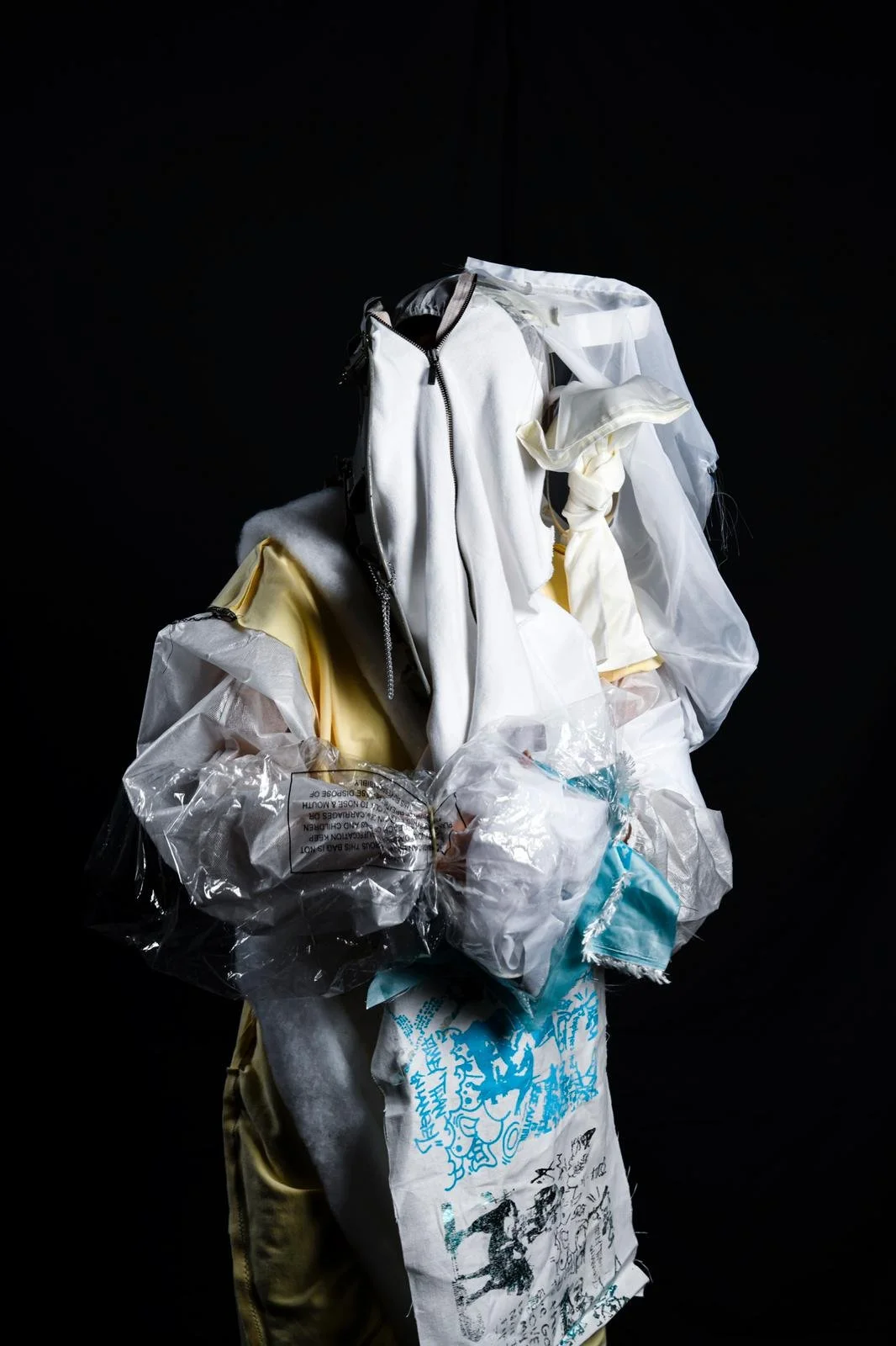 A person wearing layered protective gear, including a white hazmat suit, gloves, and a face shield, holding a plastic bag with blue and white items inside, against a black background.