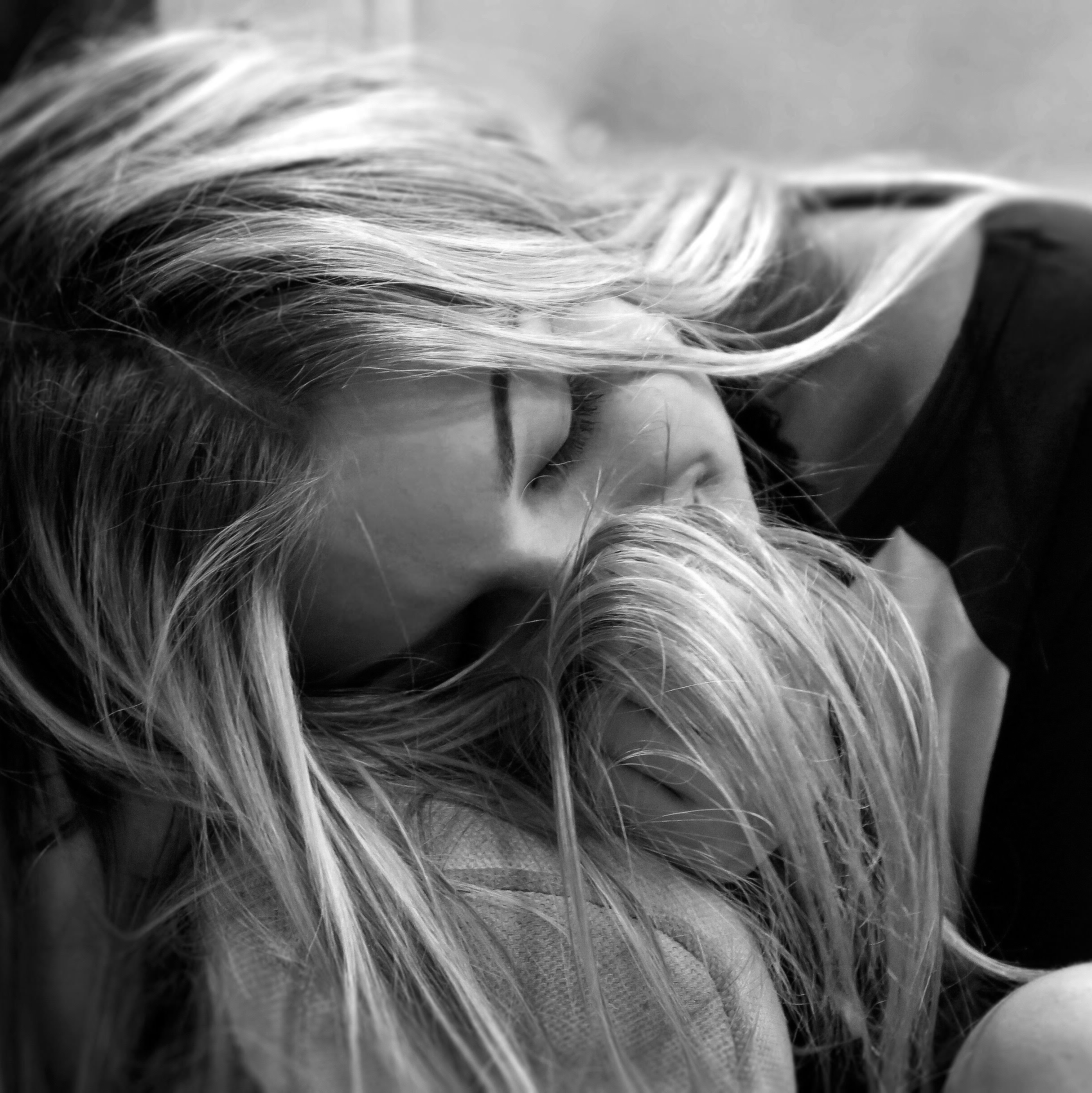 A black and white photo of a woman with long, wavy hair, resting her head on a person, with her eyes closed.