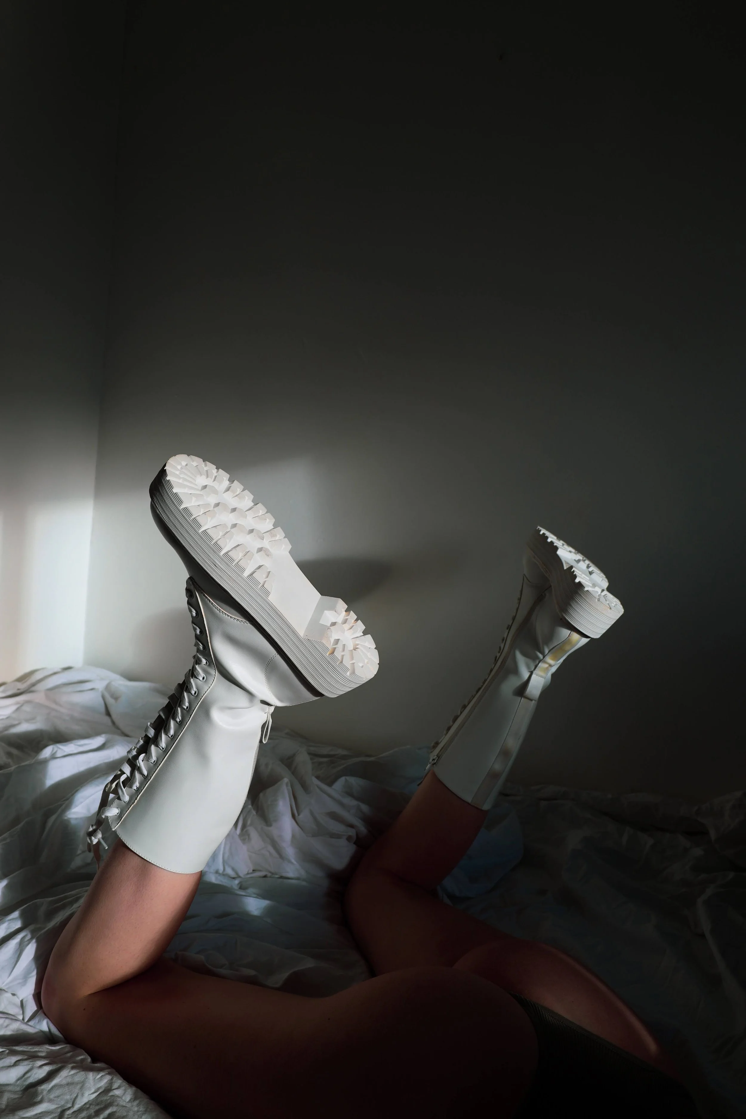 Person lying on bed with legs raised, wearing knee-high white boots with chunky soles, in a room with white bedsheets and a plain wall.