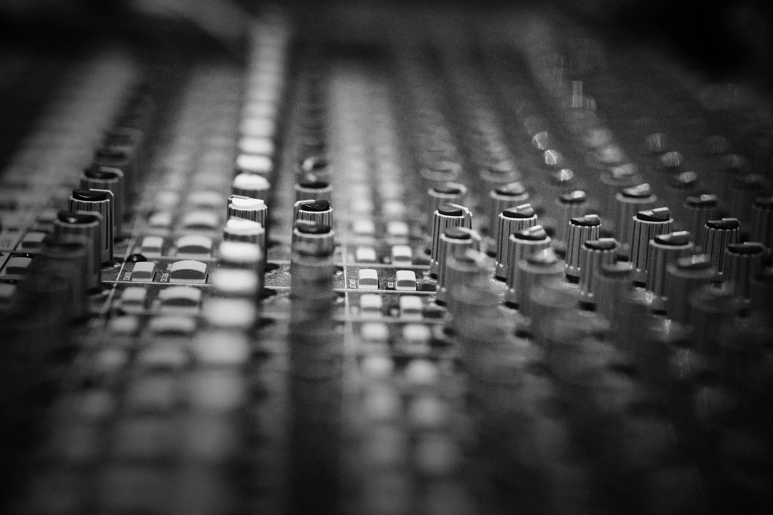 Close-up of an audio mixing console with knobs and faders in black and white.