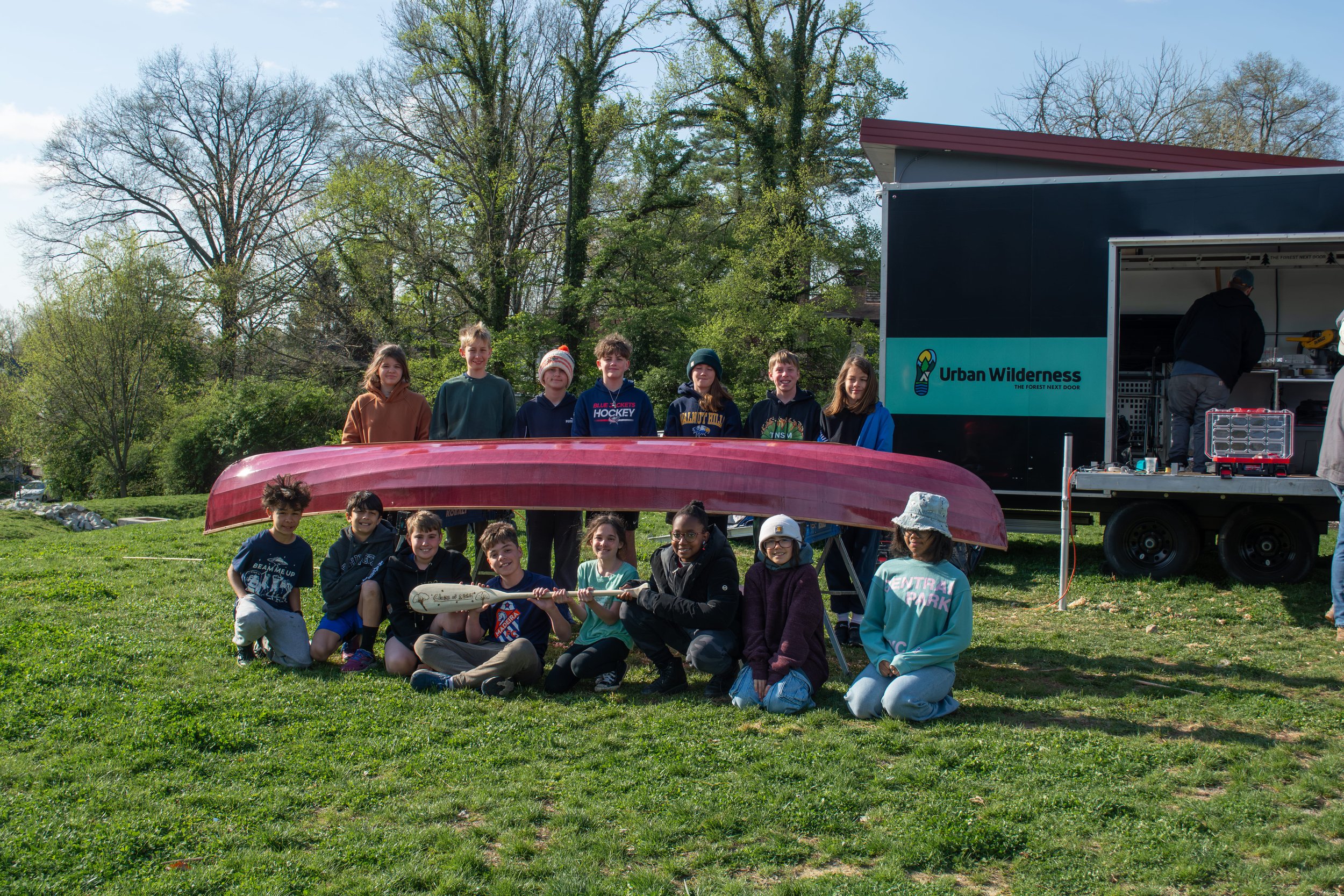 Group of children and teenagers outdoors with a canoe and a trailer labeled 'Urban Wilderness, The Forest Next Door.' The group is posing on the grass, some kneeling and some standing, in a park setting with trees and a clear sky.