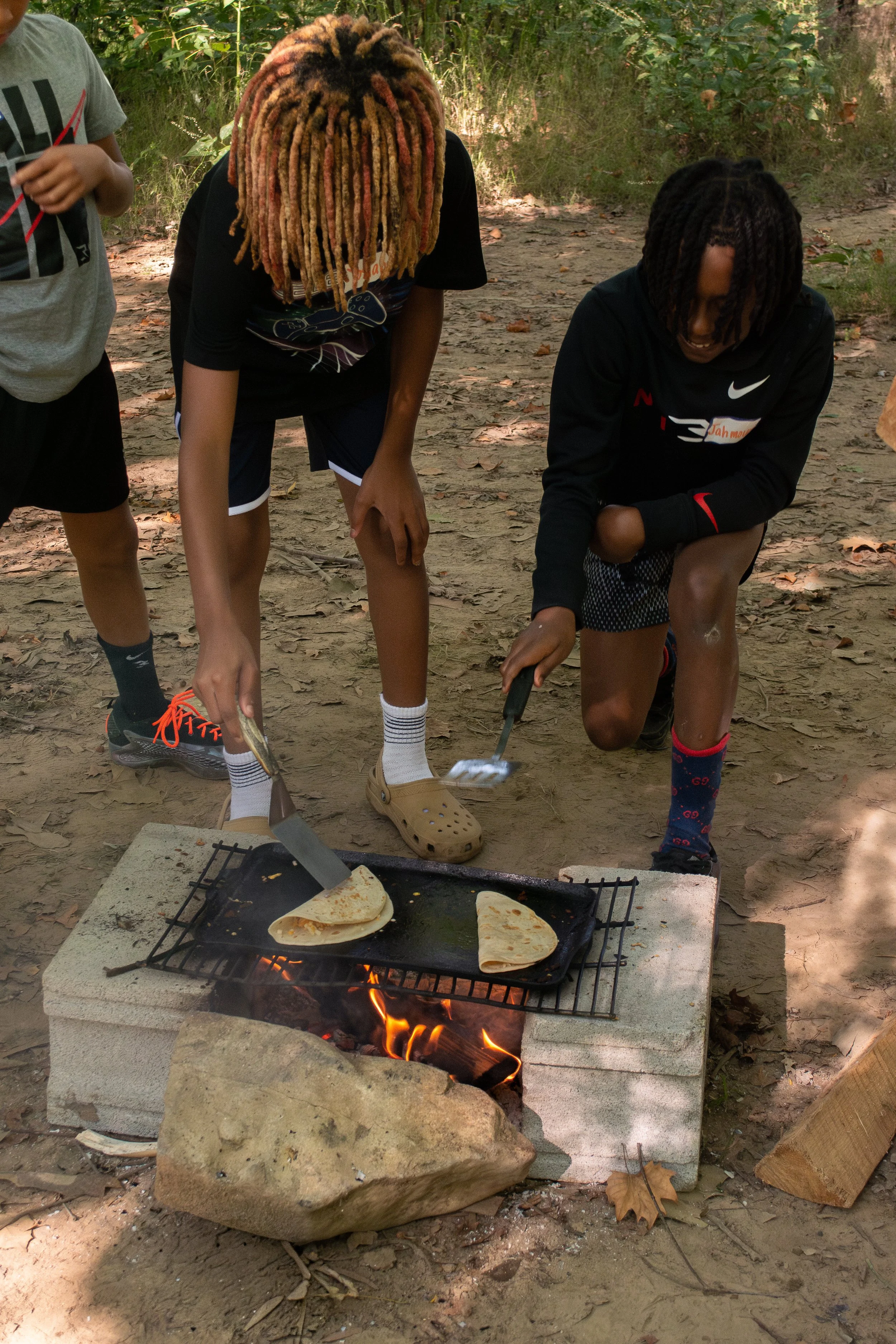 Three boys cooking flatbread on a grill outdoors in a wooded area.