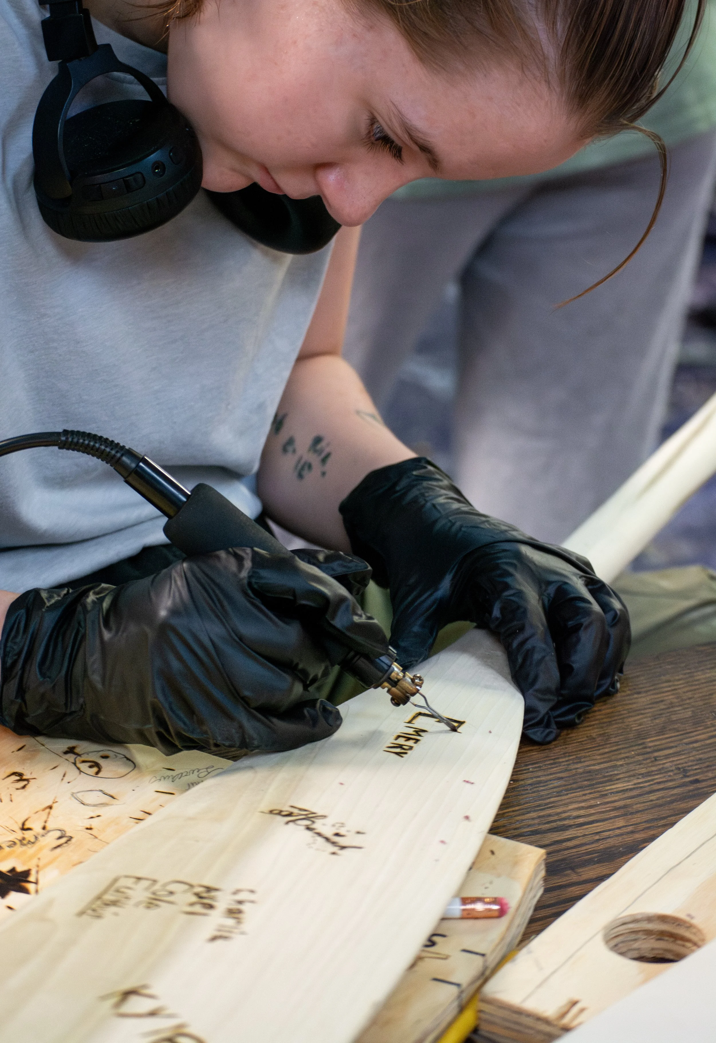 A person with light brown hair is writing in a notebook, with a tattoo on their forearm and wearing black gloves. They are using a tattoo machine or similar tool on a piece of wood with handwritten words. The person is also wearing black headphones.