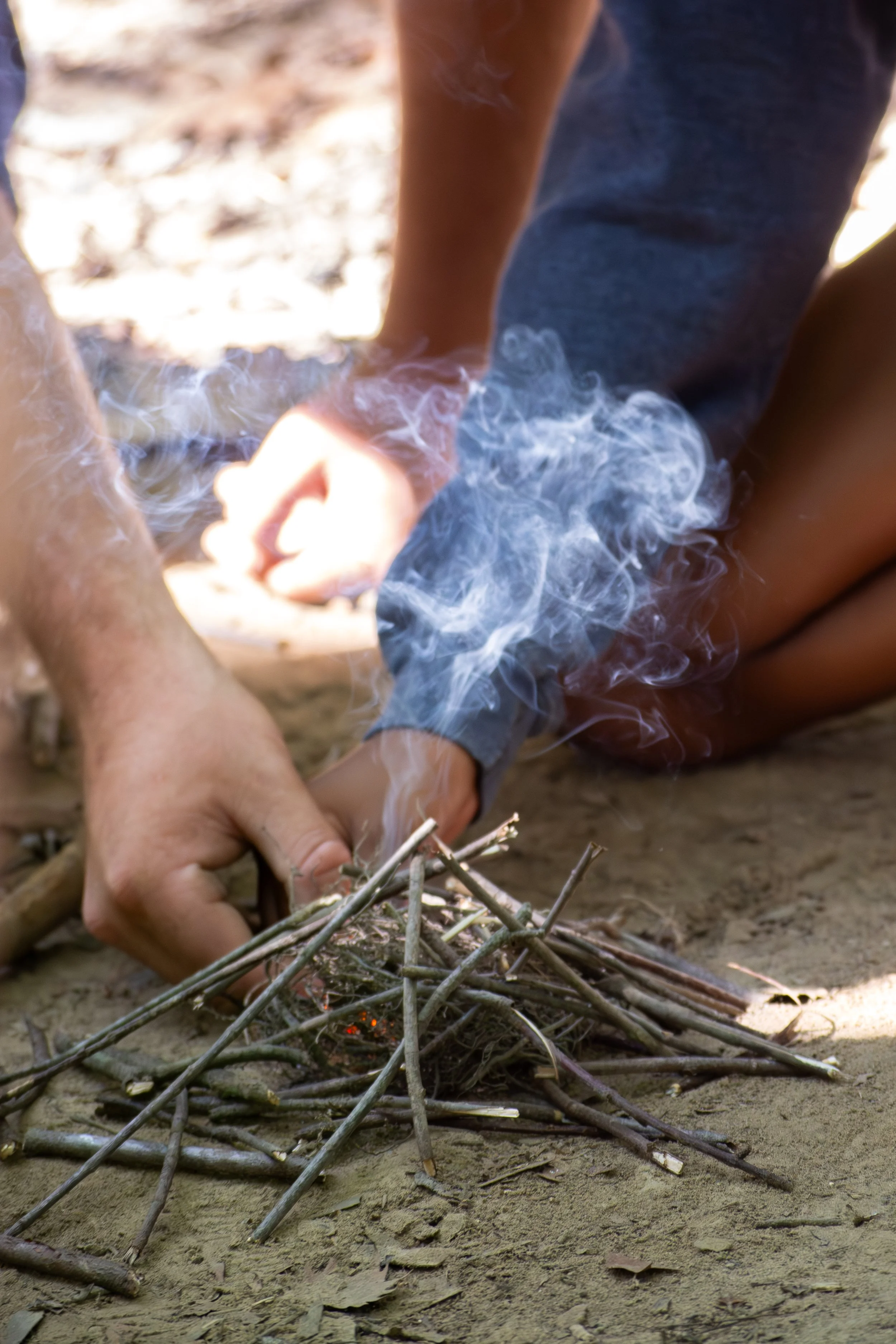 Person sitting on the ground starting a campfire with small twigs, surrounded by dry leaves and soil.